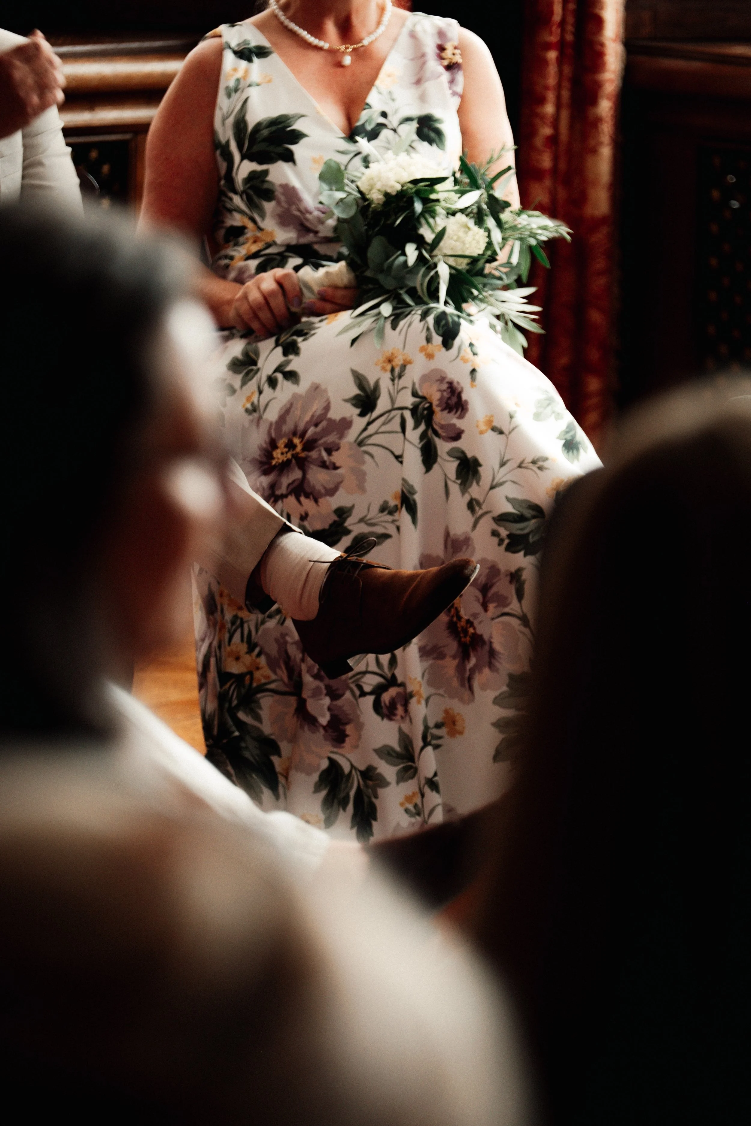 Close-up van een vrouw in een bloemrijke jurk die een boeket met witte bloemen vasthoudt, zittend met een parelketting op een warme, sfeervolle achtergrond. bruid bruiloft Arnhem kasteel