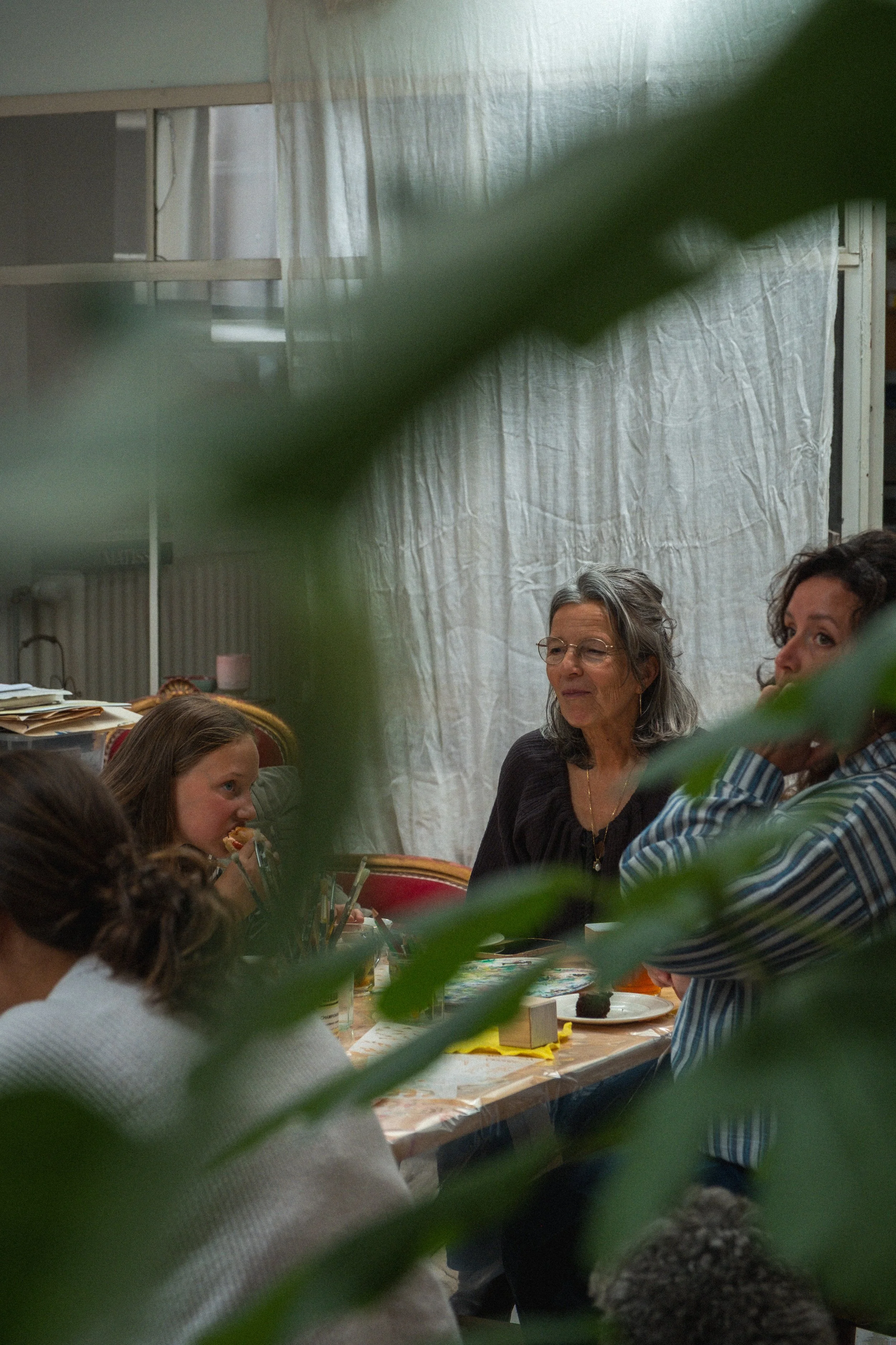 Een groep mensen zit rond een tafel tijdens een bijeenkomst of feestje, met een witte gordijn en een boekenkast op de achtergrond, gedeeltelijk verborgen door groene bladeren.