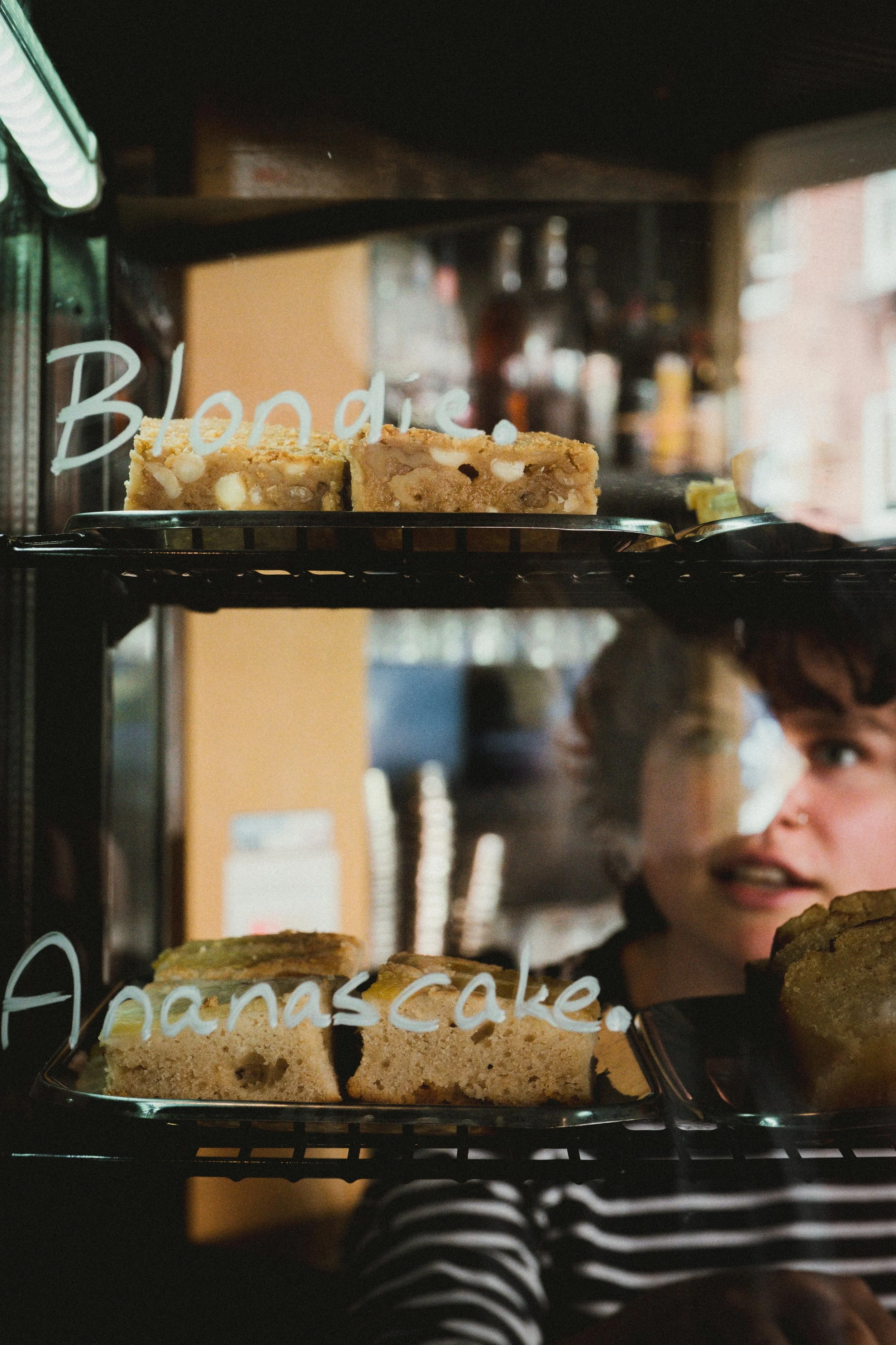 Twee stukken bananen cake in een display case met witte tekst die 'Banana cake' zegt, en een persoon met een zwart-wit gestreept shirt die ernaast staat.