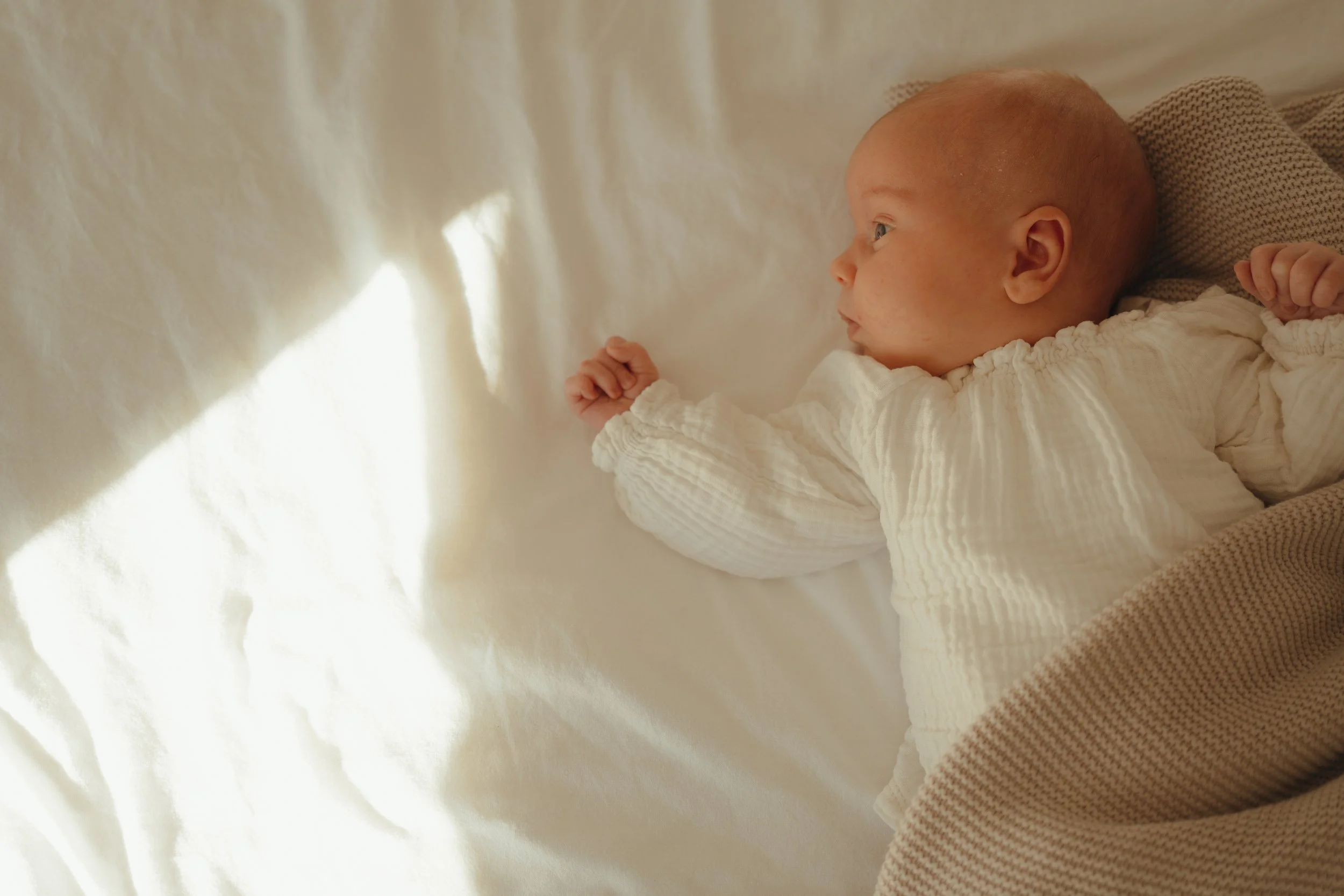 Babyliefje ligt op een witte deken, gekleed in een rompertje en bedekt met een bruin gestreept dekentje. Kijkend naar buiten, met licht dat door een gordijn schijnt. newborn baby 