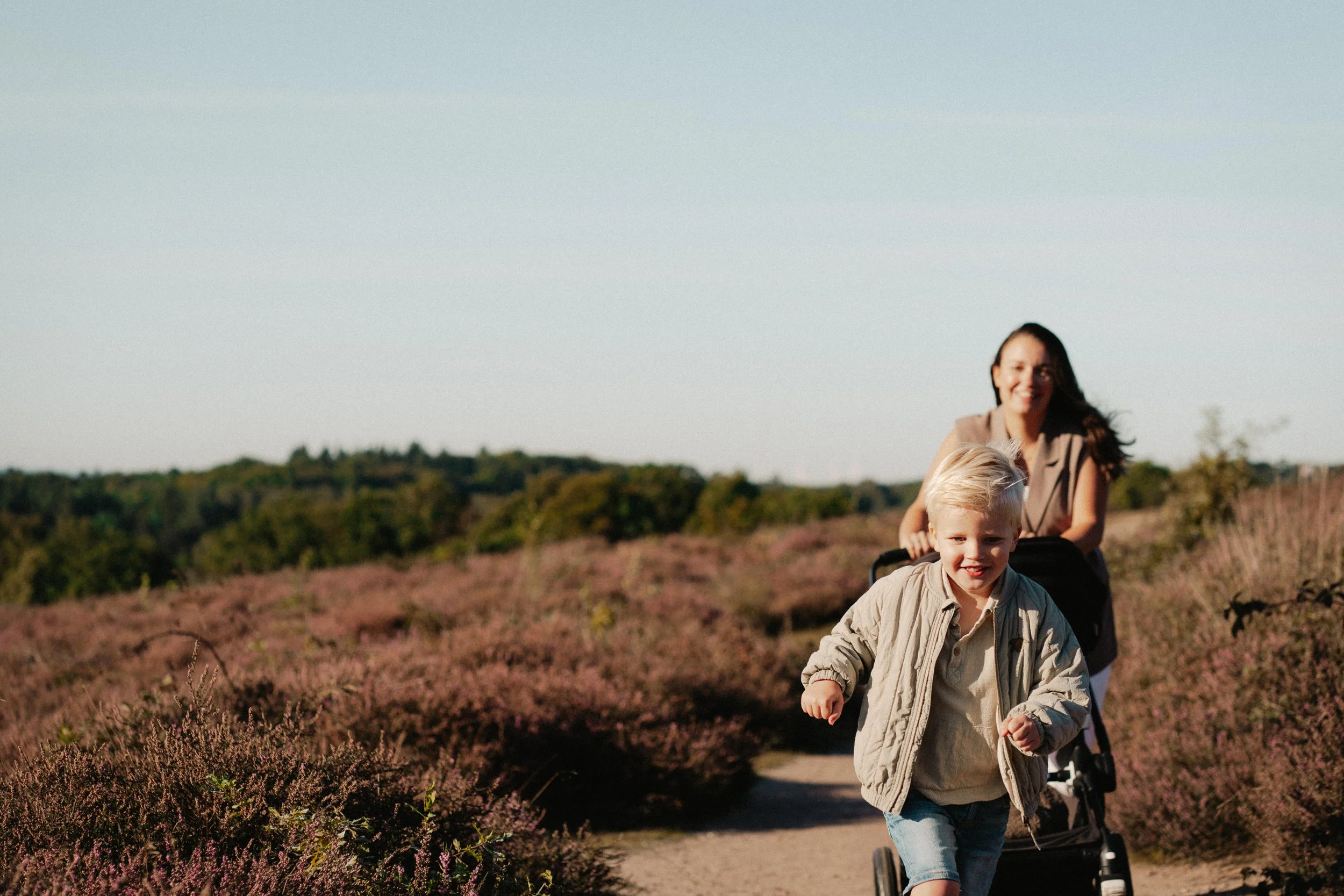 Een vrouw en een kind lopen samen door een veld met bloemen, de vrouw duwt een kinderwagen, het kind is vooraan en glimlacht.