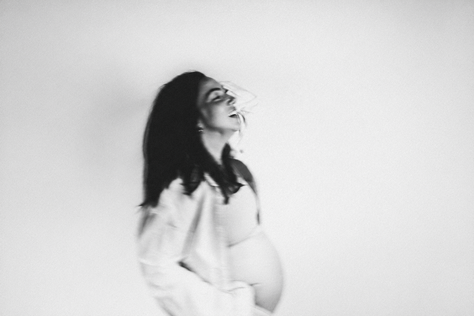 Zwarte en witte foto van een zwangere vrouw met lang zwart haar, lachend en met haar hand op haar buik, tegen een effen lichte achtergrond. vogue magazine blurry