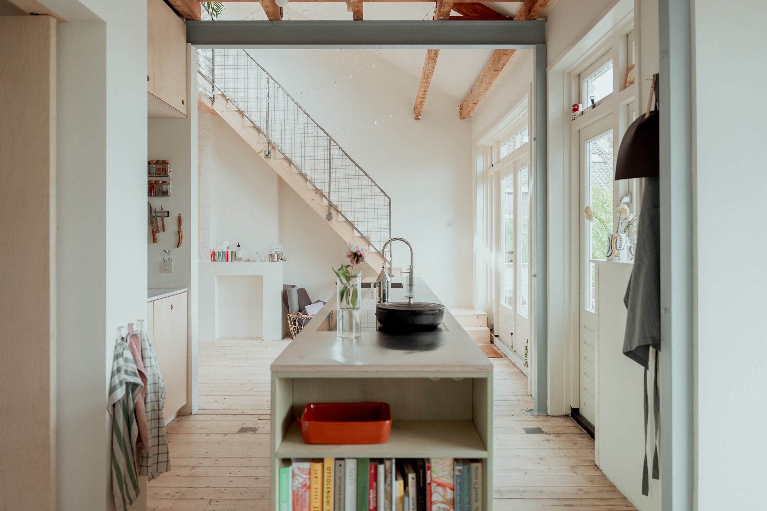 Een open keuken met een eiland, witte en houten keukenapparatuur, en grote ramen die veel natuurlijk licht binnenlaten in een modern huis met houten vloer en zichtbare houten plafondbalken.