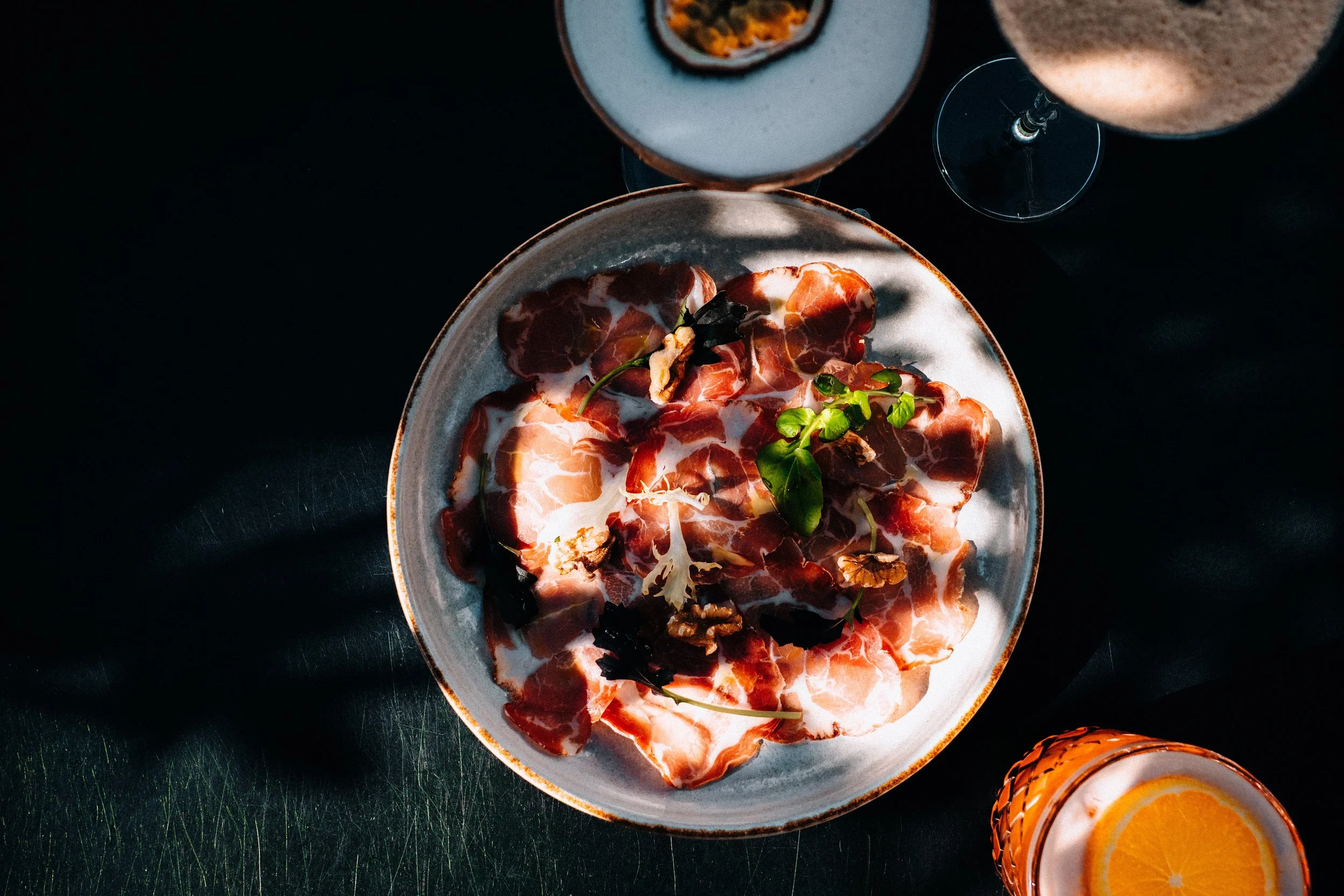 Een bijna lege kom met dun gesneden prosciutto, garnering met microgroen en noten, naast twee glazen met een drankje met ijs en een schijfje sinaasappel, op een zwarte tafel. pornstar martini horeca fotografie Arnhem