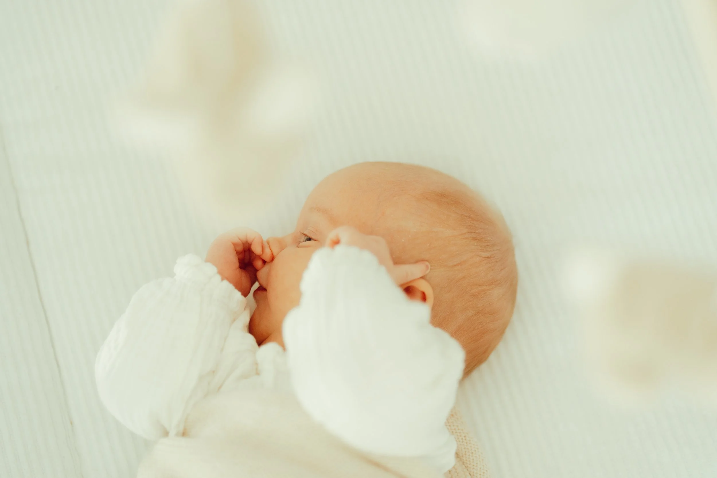 Een pasgeboren baby ligt op een witte bedje en zuigt aan haar duim.