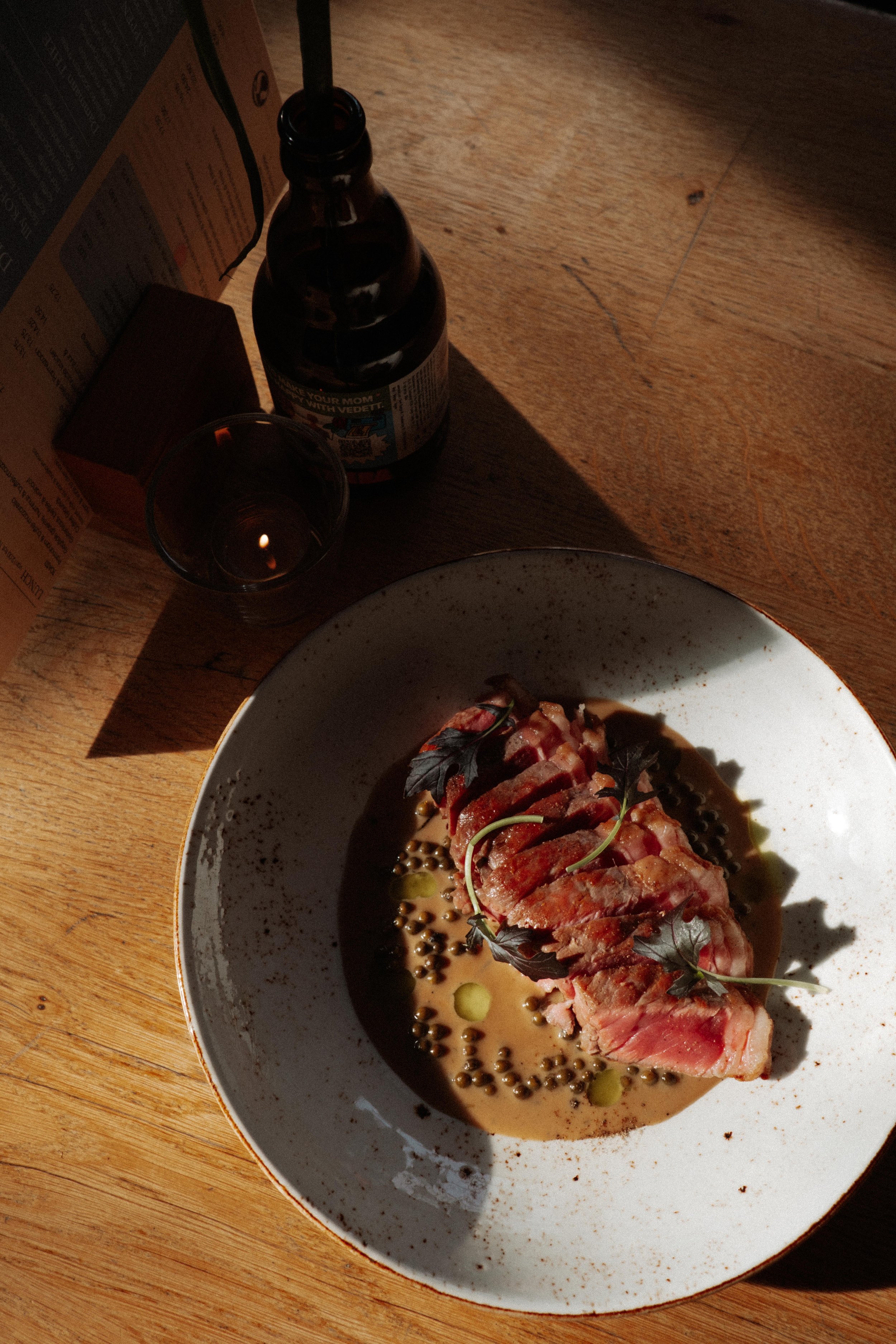 Een bord met een stuk vlees in saus, garnering met kruiden en olijfolie, op een houten tafel naast een glas donkere drank en een fles bier.