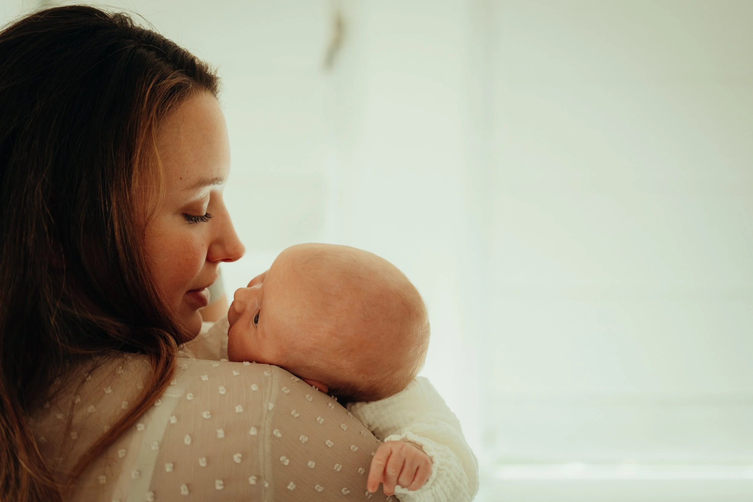 Een vrouw met bruin haar knuffelt een baby die naar haar kijkt en haar aanraakt, tegen een lichte achtergrond.