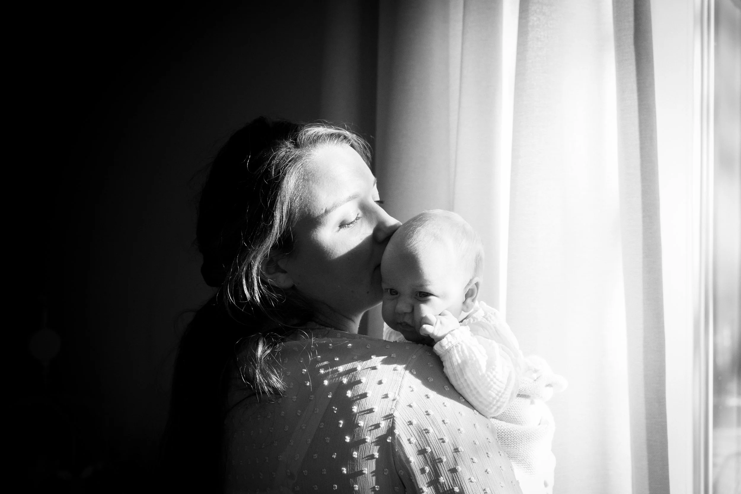 Zwart-wit foto van een vrouw die een baby koestert bij een raam met gordijnen, beide gezichten naar de ramen gericht.