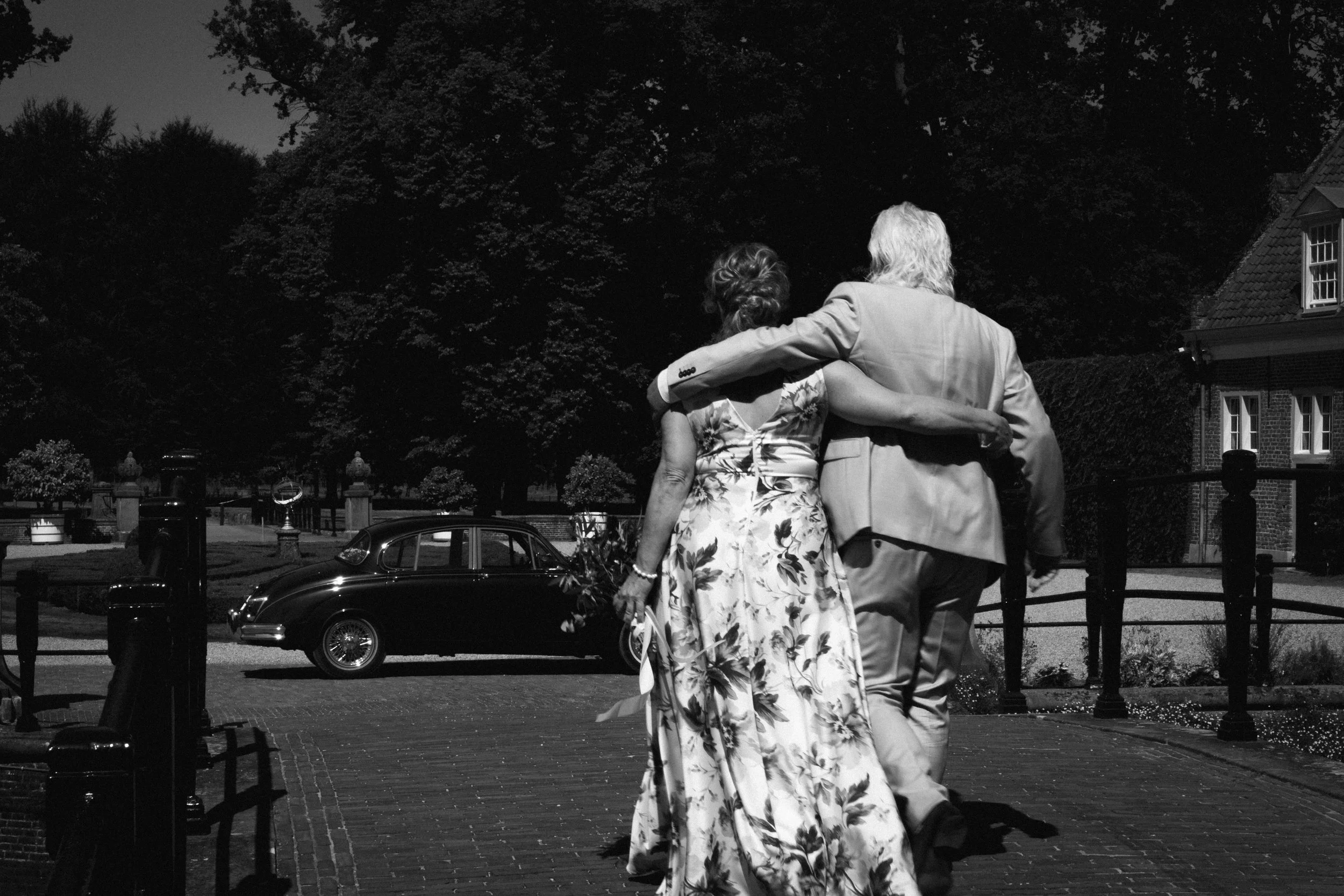 Een zwart-wit foto van twee mensen die samen wandelen, één man met grijs haar en een vrouw in een jurk met bloemen, in een tuin met bomen en een huis op de achtergrond.