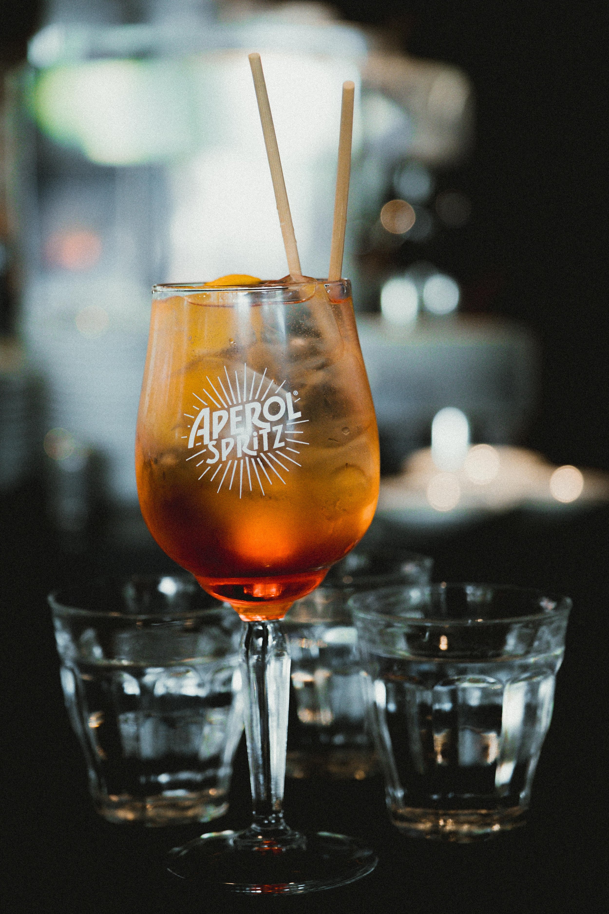 Een close-up van een Aperol Spritz cocktail in een wijnglas met ijs, een schijfje sinaasappel en twee rietjes, geplaatst op een glas met ijs. De achtergrond is wazig met lichte vlekken.