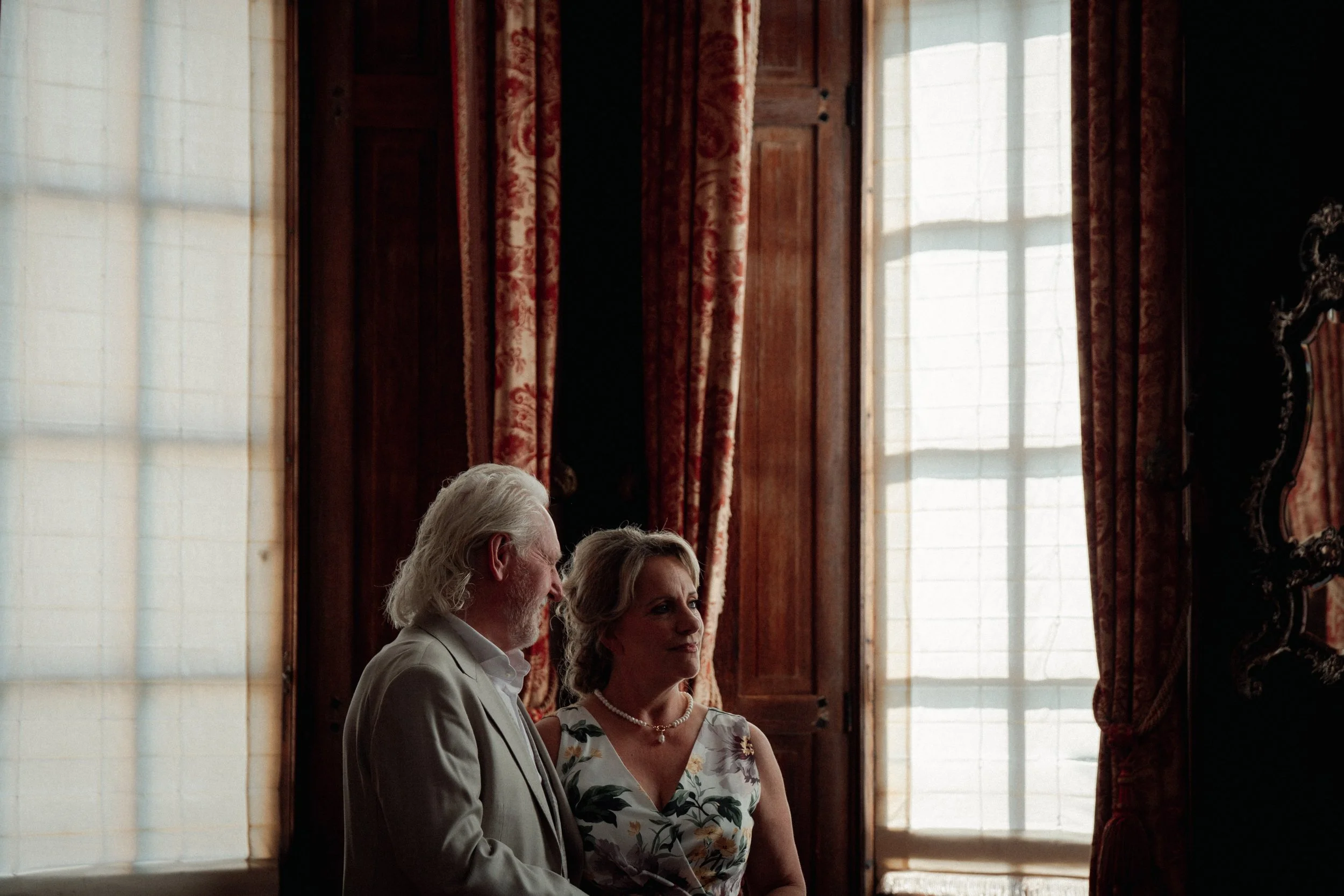 Een oudere man en vrouw zitten naast elkaar in een kamer met grote ramen en rode gordijnen tijdens een formele gelegenheid. bruiloft bruidspaar ceremonie