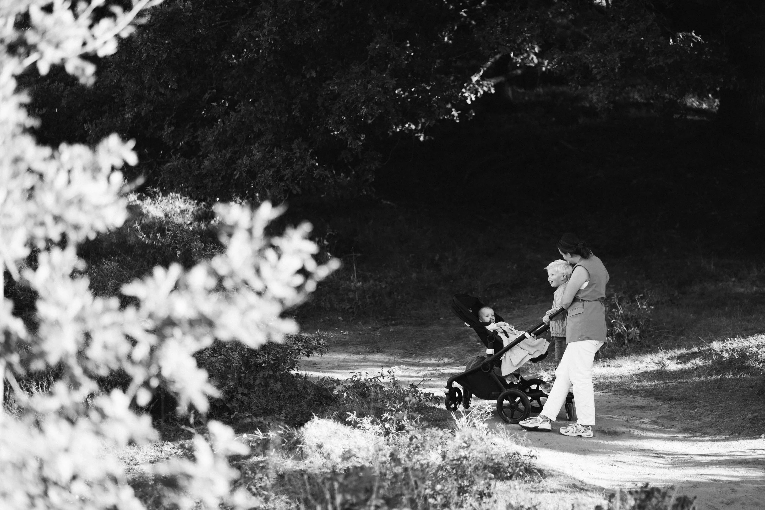 Een vrouw die met twee jonge kinderen wandelt in een park of bosgebied. Eén kind zit in een kinderwagen, terwijl de vrouw met het andere kind naast haar loopt.