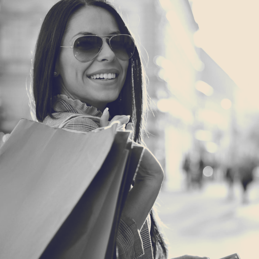 Woman wearing sunglasses and smiling while holding shopping bags in an outdoor city setting.