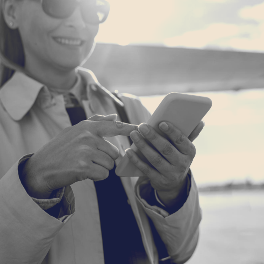 A woman with glasses smiling and using a smartphone outdoors.