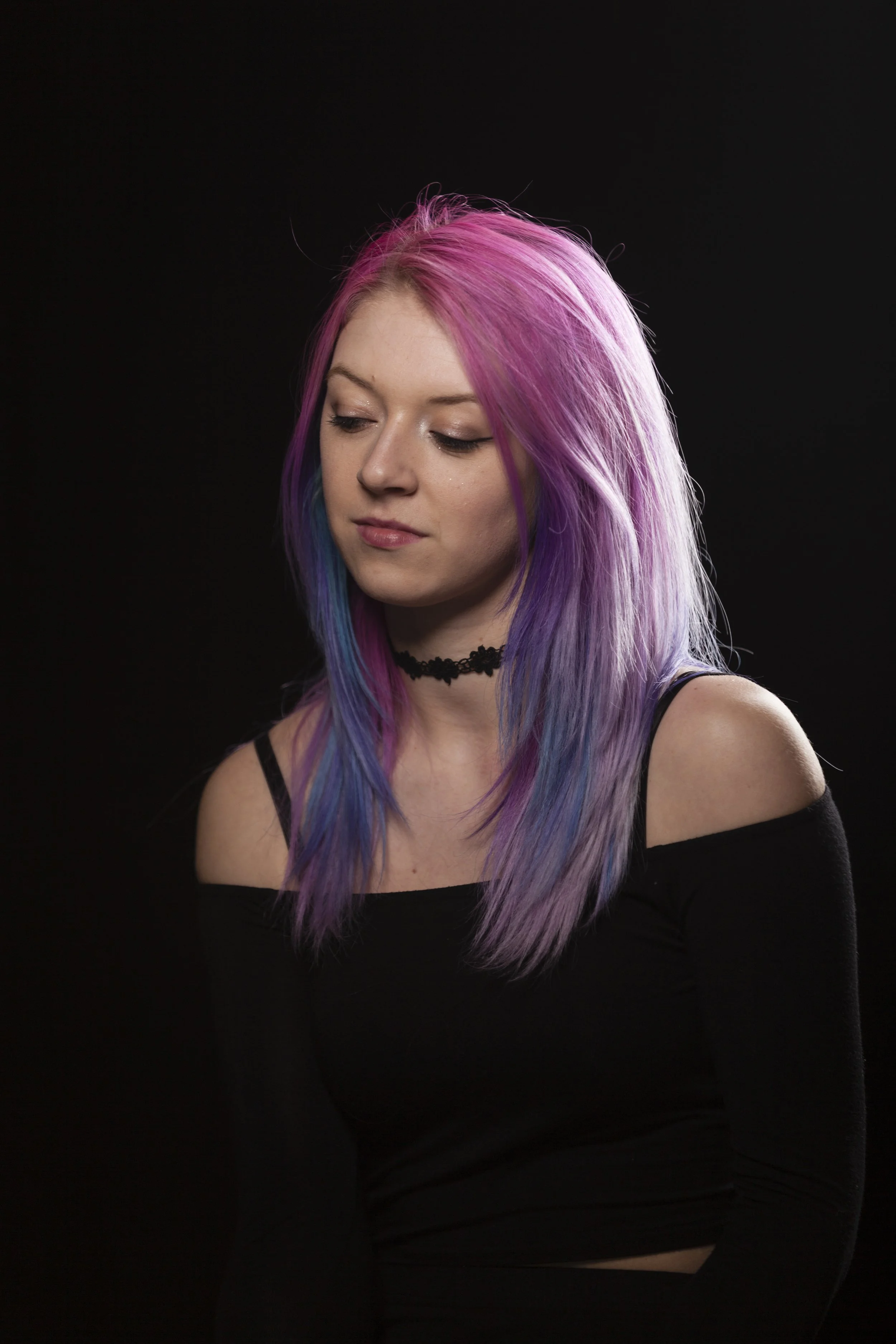 A young woman with long, straight hair dyed in pink, purple, and blue hues. She is wearing a black off-the-shoulder top and a black choker necklace. Her eyes are closed, and she is looking slightly downward against a plain black background.