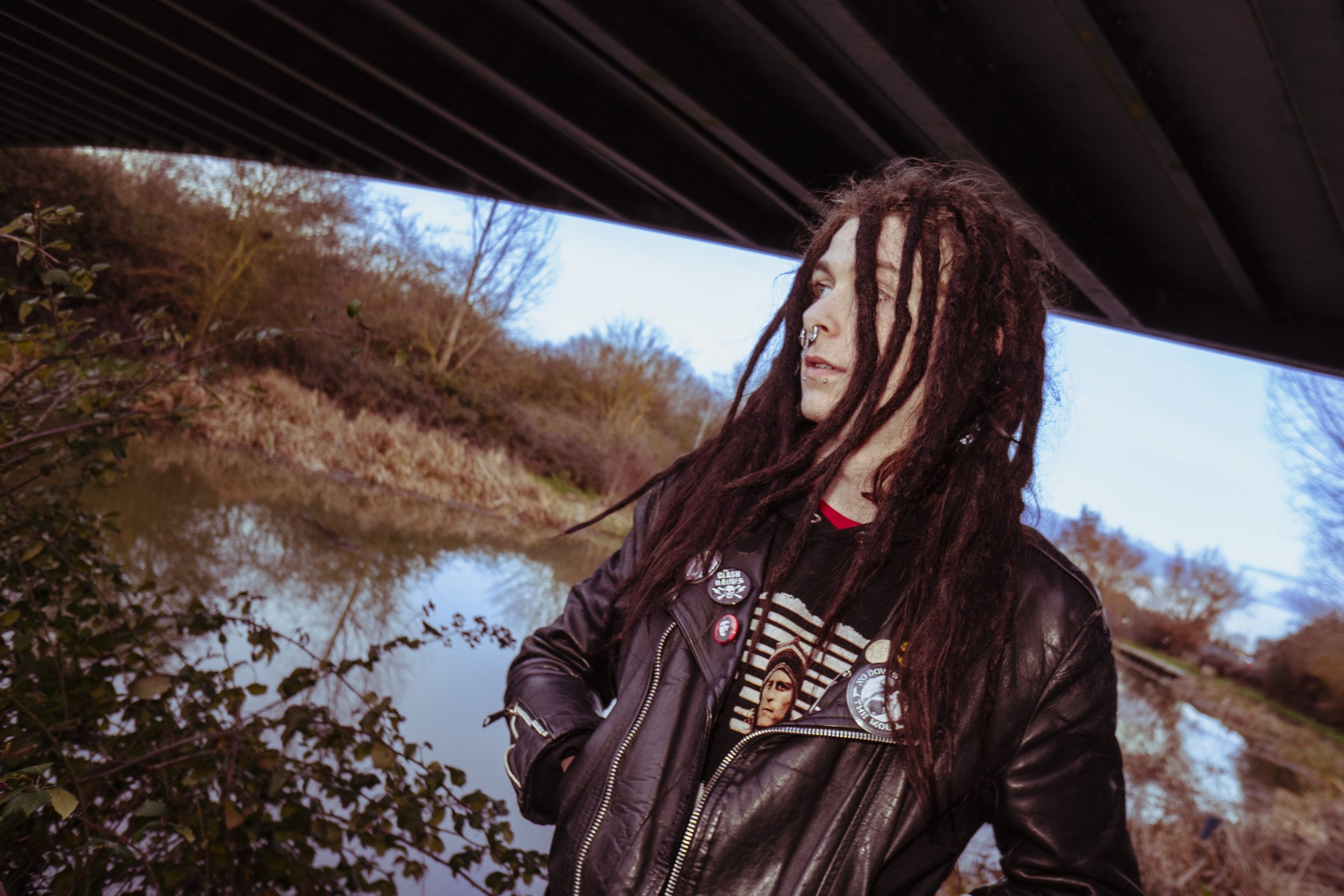 A person with long dreadlocks, wearing a black leather jacket with patches, stands near a river with trees in autumn colors under a bridge.