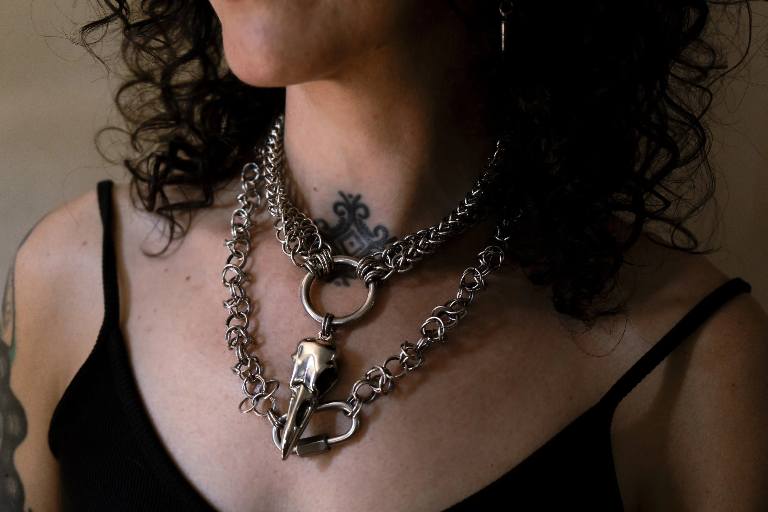 Close-up of a person's neck and chest area with multiple silver necklaces, one of which has a large ring and a skull pendant, and a tattoo partially visible at the base of the neck.