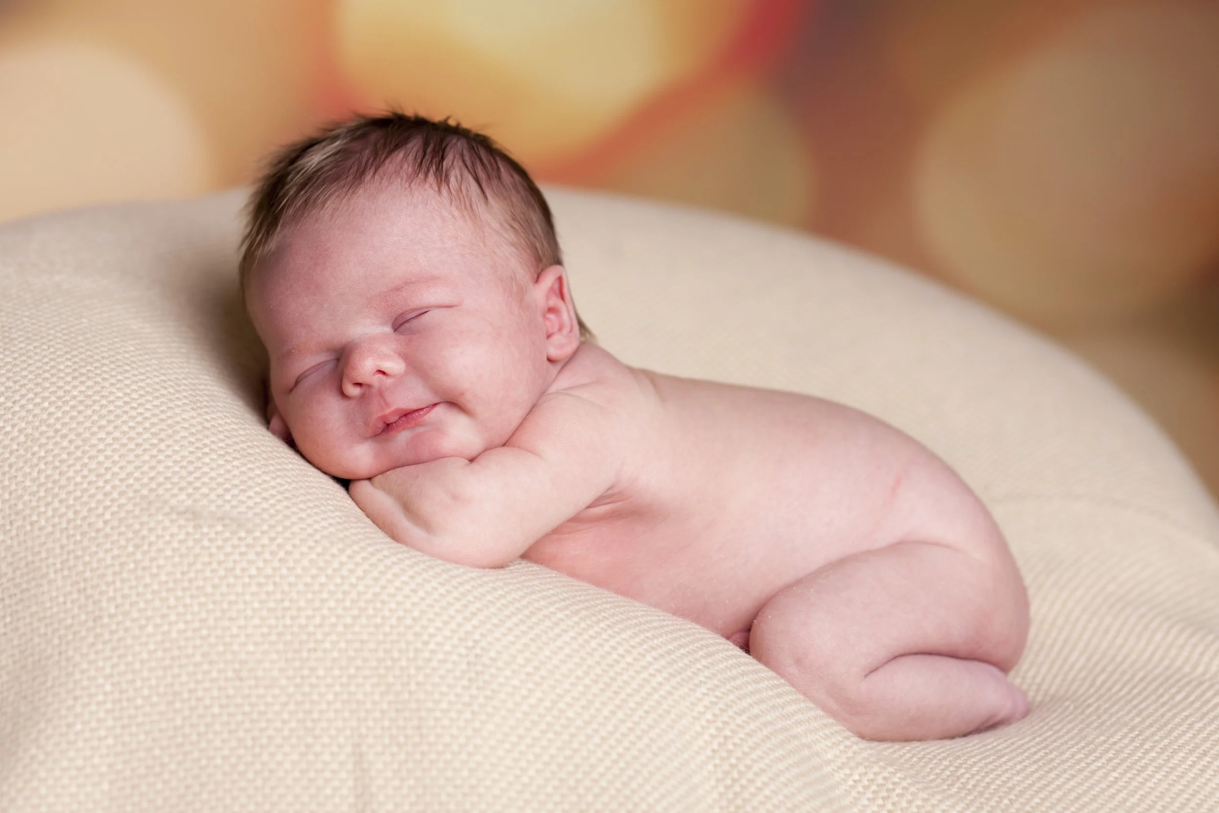 A sleeping newborn baby lying on a soft, beige surface with eyes closed and a slight smile, showcasing tiny features and delicate skin.
