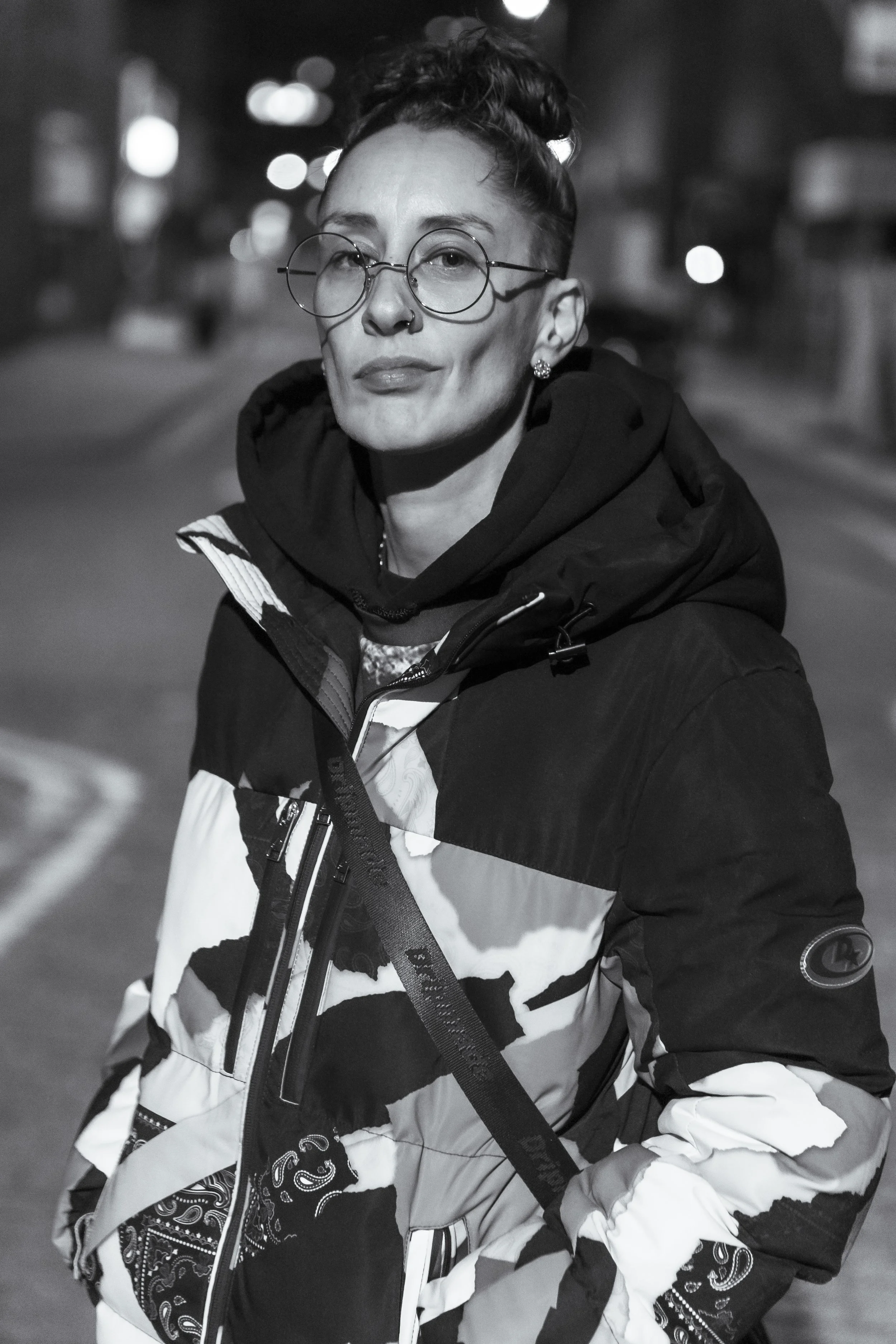 A woman with short hair, wearing glasses and earrings, dressed in a striped jacket with a paisley pattern, standing outdoors at night with blurred city lights in the background.