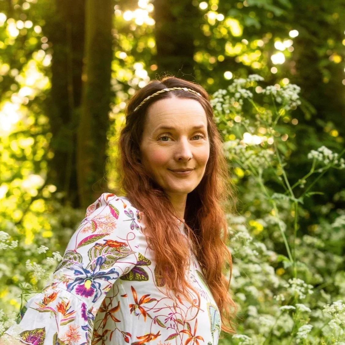 A woman with long red hair and a headband, smiling in a lush green forest.