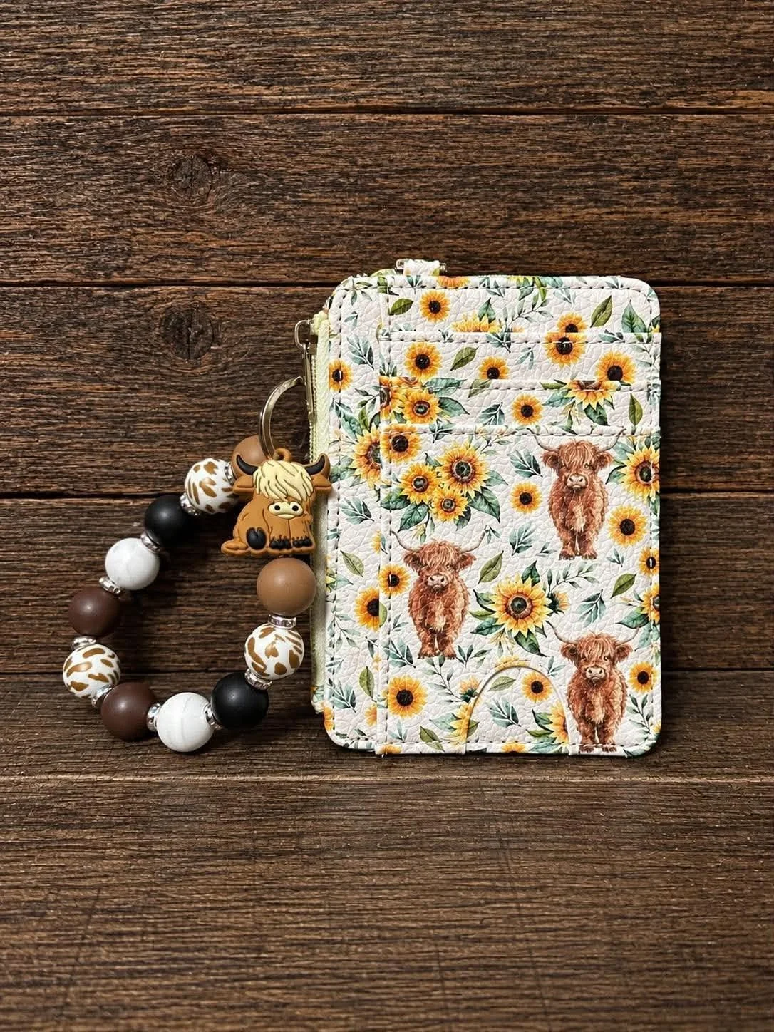 A fabric folder with a sunflower and cow pattern is placed on a wooden surface with a beaded keychain attached.