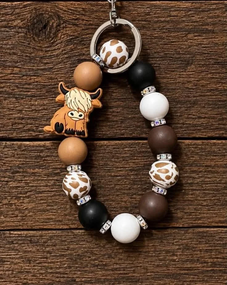 A beaded keychain with a llama-shaped silicone figure, alternating brown, black, white, and animal print beads, attached to a metal ring on a wooden surface.