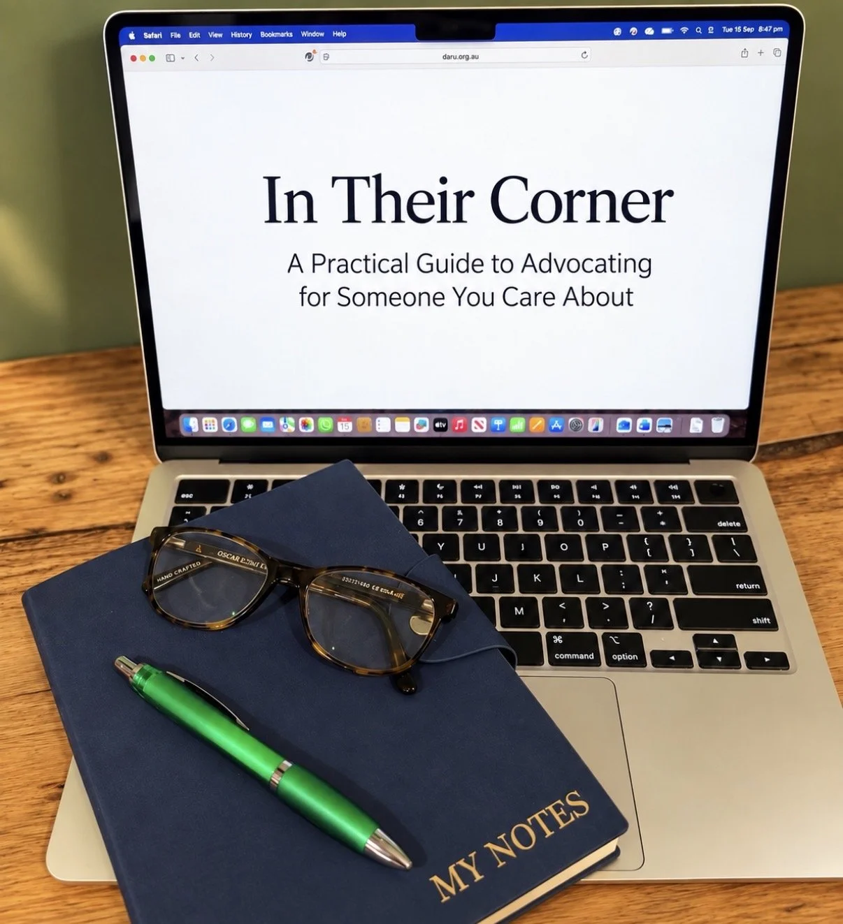 A laptop with a webpage titled 'In Their Corner' on the screen, a pair of glasses, a green pen, and a blue notebook labeled 'My Notes' resting on a wooden table.