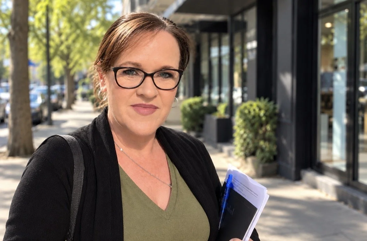 A woman with brown hair, glasses, and a slight smile standing on a city sidewalk with trees and storefronts behind her, holding a notebook and pen.