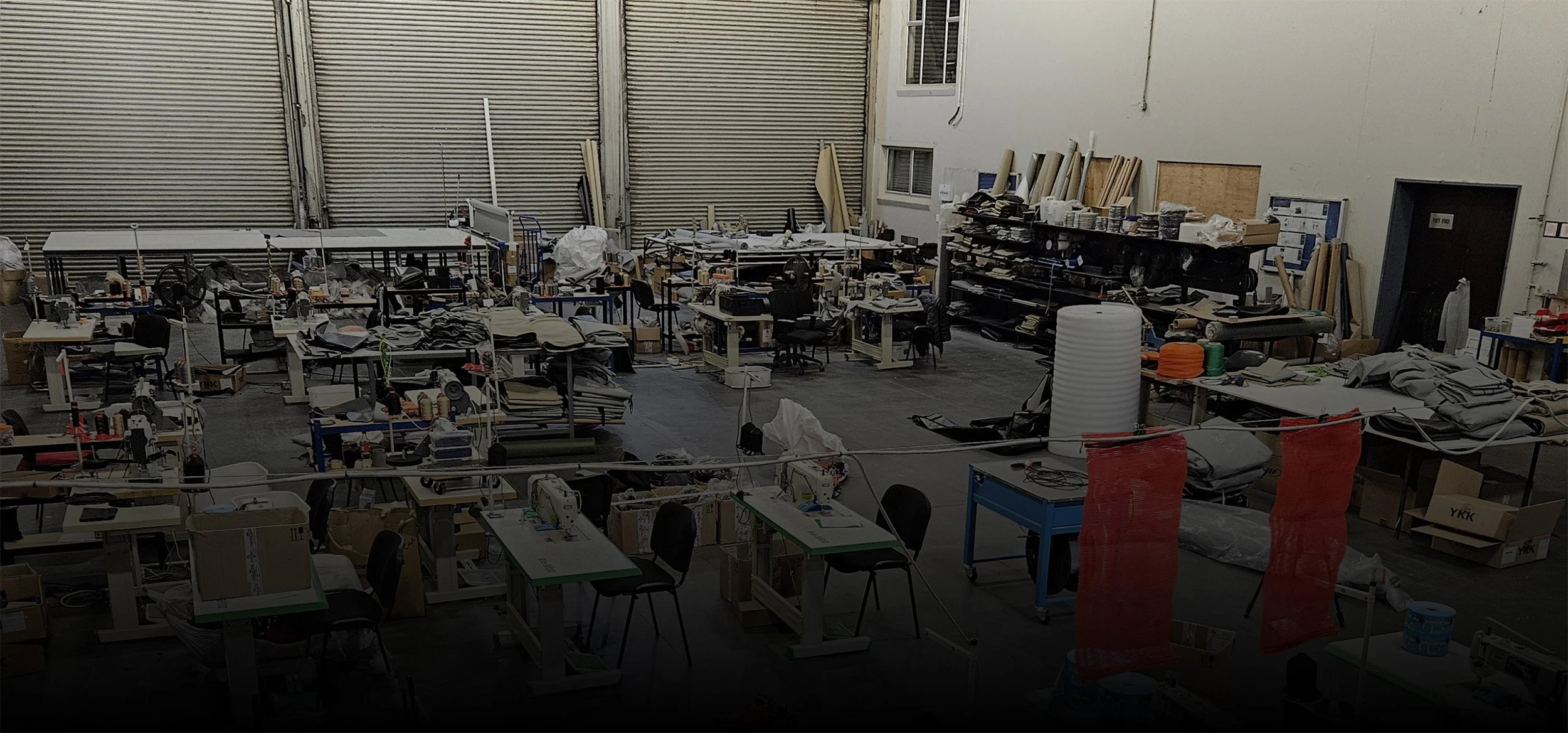 A factory or workshop filled with desks, chairs, and various equipment, with supplies and materials scattered throughout the space.