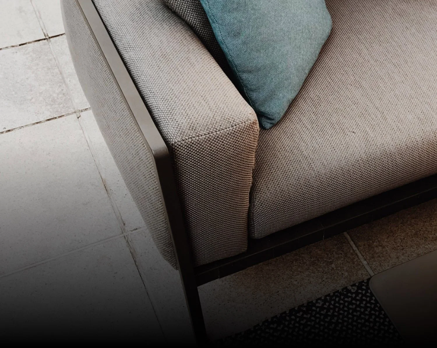 Close-up of a beige upholstered sofa with teal and beige cushions, located next to tiled flooring and a dark brown baseboard.