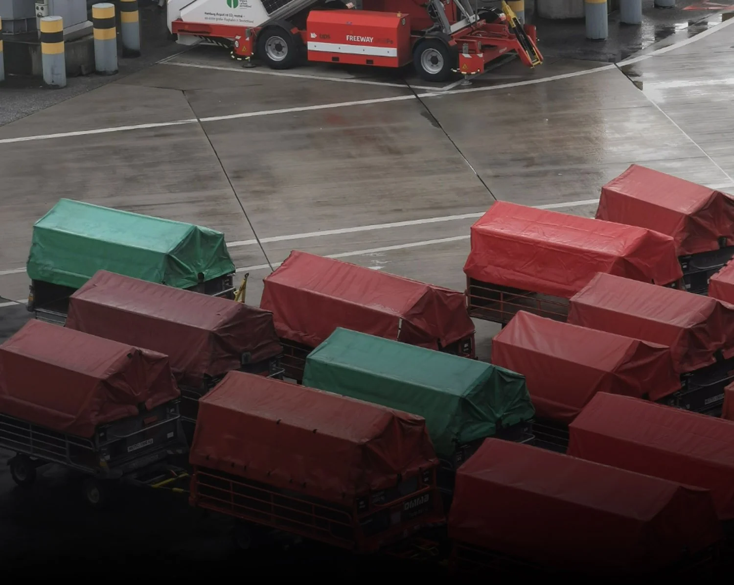 Several luggage carts with red and green covers parked at an airport gate, wet tarmac in the background.