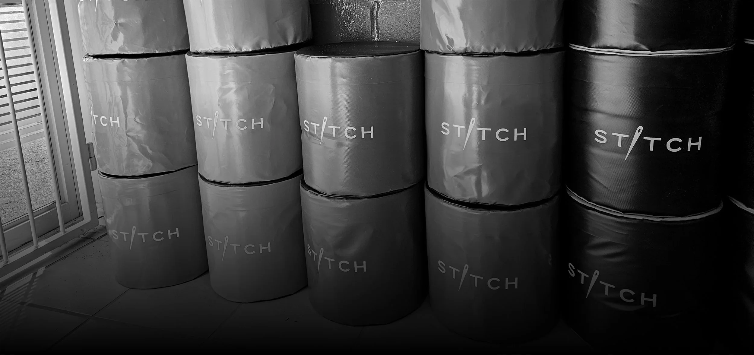 Stacked soft play cylinders with the Stitch SA logo printed on them, stored against a wall near a sliding glass door with metal bars.