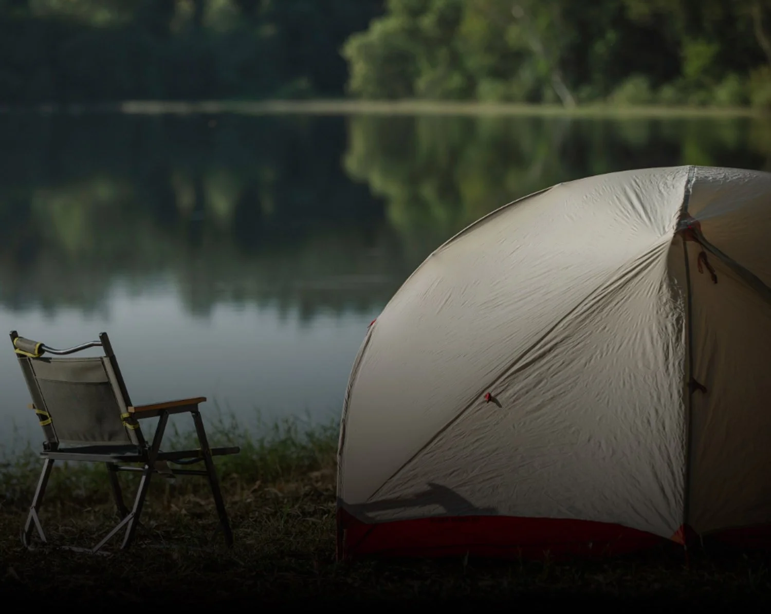 A camping tent and a camping chair set up by a lakeshore, with trees reflected in the water in the background.
