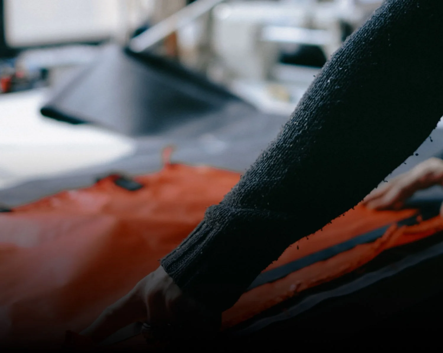 Close-up of a person's arm and hand with a black sleeve, working on an orange piece of fabric gear, with a blurred background of a white surface and objects.