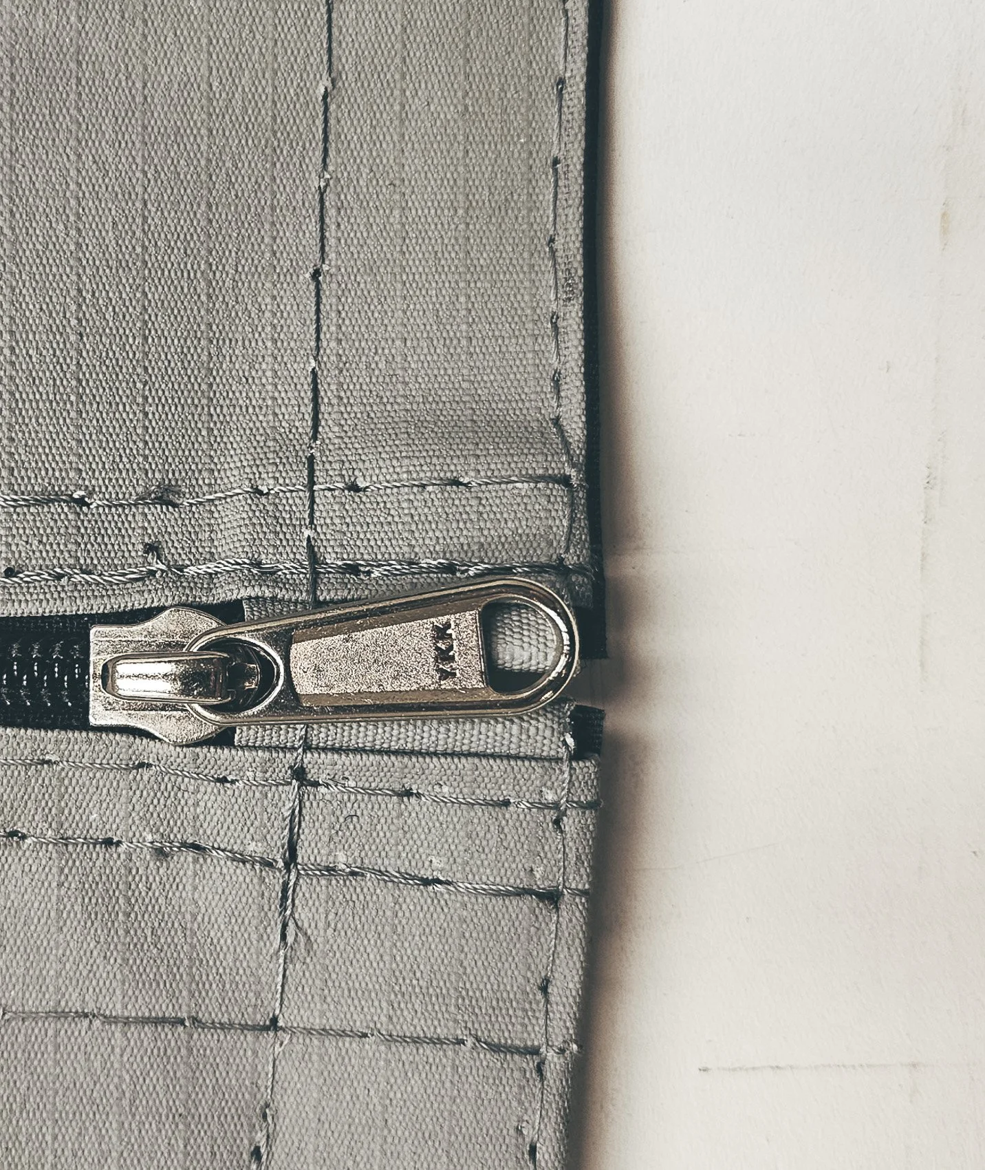 Close-up of a silver zipper on a gray fabric with visible stitching, next to a plain white wall.