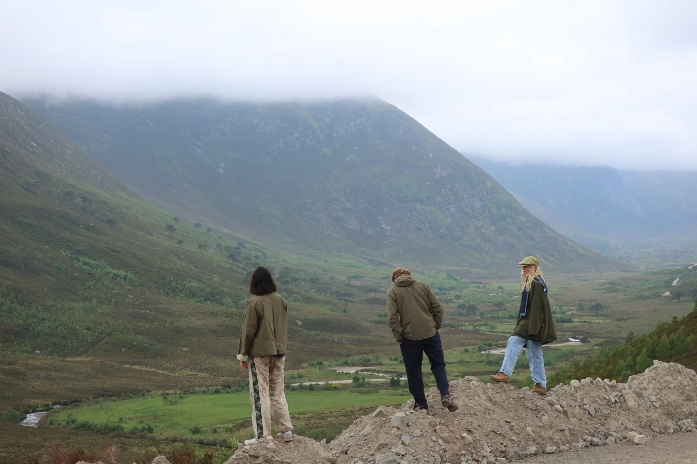 A group of four people standing outdoors on rocky ground, overlooking a lush green valley with mountains partially covered in fog in the background on a cloudy day.