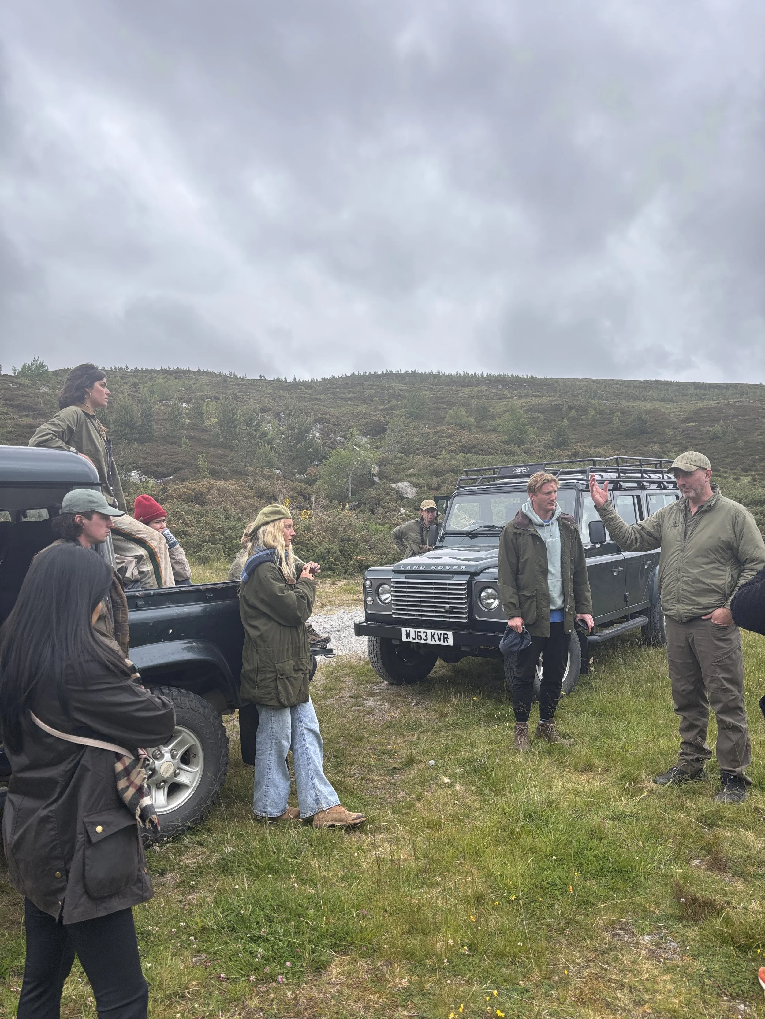 Group of people outdoors in a grassy area with Land Rover vehicles, discussing under cloudy sky, hillside in background.