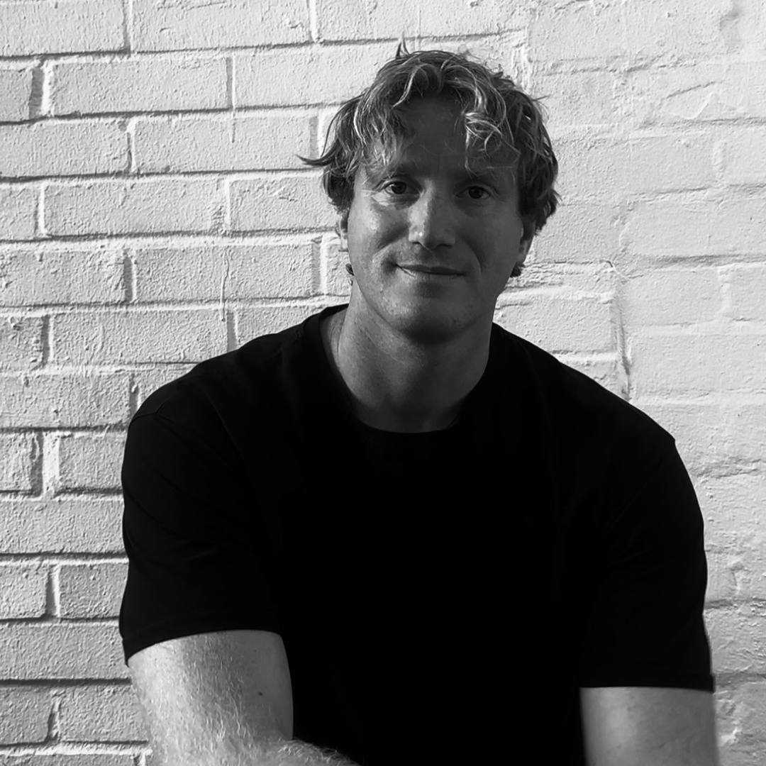 A black and white photo of a young man with wavy hair, wearing a dark t-shirt, sitting in front of a brick wall.