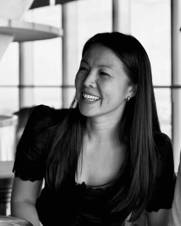 A woman with long dark hair smiling and looking to the side in a modern building with large windows.