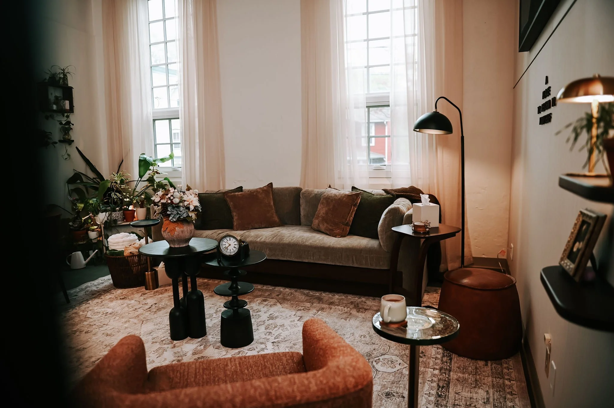 A cozy living room with a beige sofa, various pillows, potted plants, and a coffee table with decorative items, all illuminated by natural light from large windows with sheer curtains.