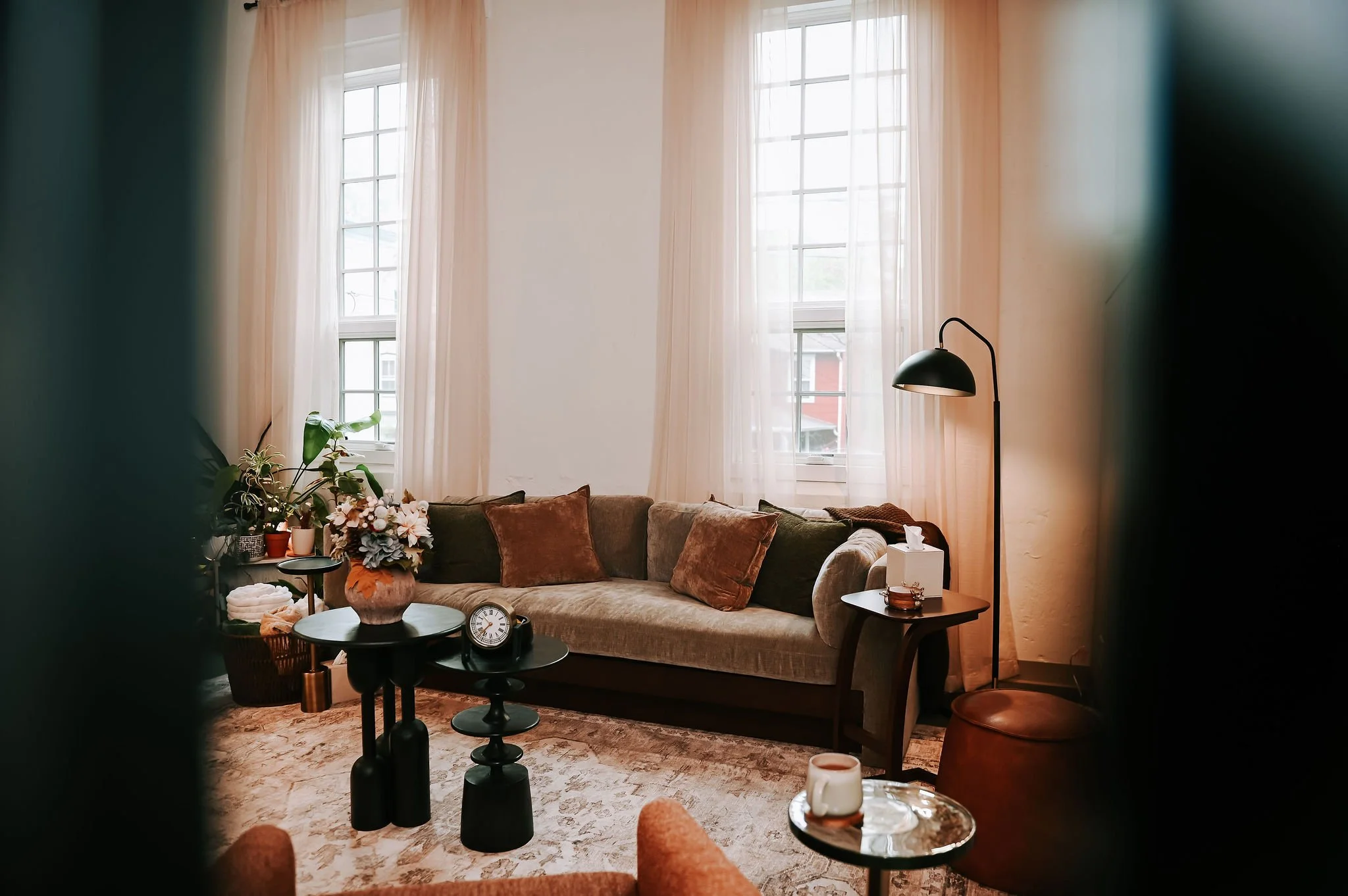 Living room with large windows, sheer curtains, beige sofa with brown throw pillows, black floor lamp, flowers on a side table, and a patterned rug.