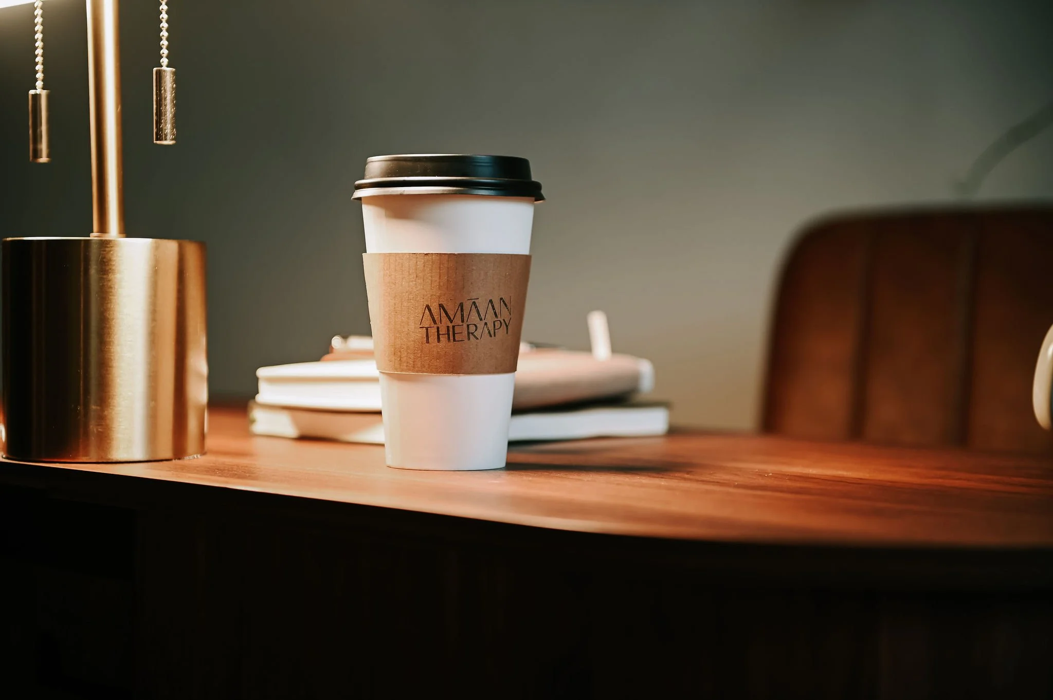A paper coffee cup with a black lid and a brown sleeve that says 'AMAM THERAPY' on a wooden table, with stacked books in the background and a brass lamp on the left.