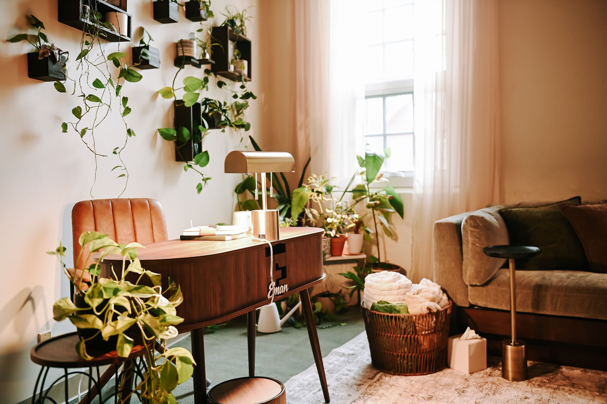 A cozy living room with a wall decorated with black shelves holding potted plants, sunlight streaming through large windows with sheer curtains, a wooden sideboard with books and a metallic lamp, a beige tufted sofa, and various plants and baskets ad