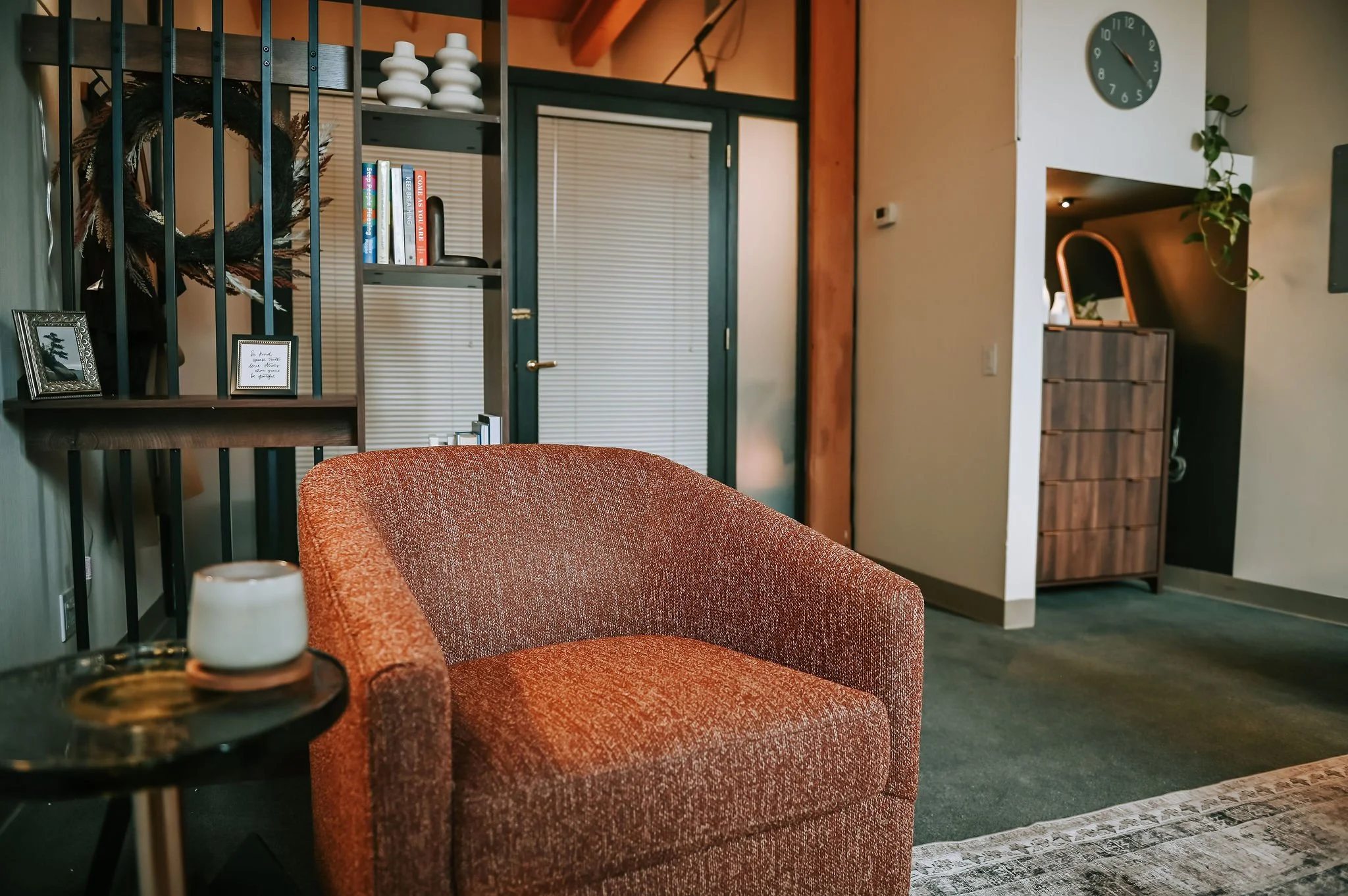 Living room with orange chair, black side table, and decorative items. Background features bookcase with books, wreath, and framed photos, along with doorway and clock on wall.