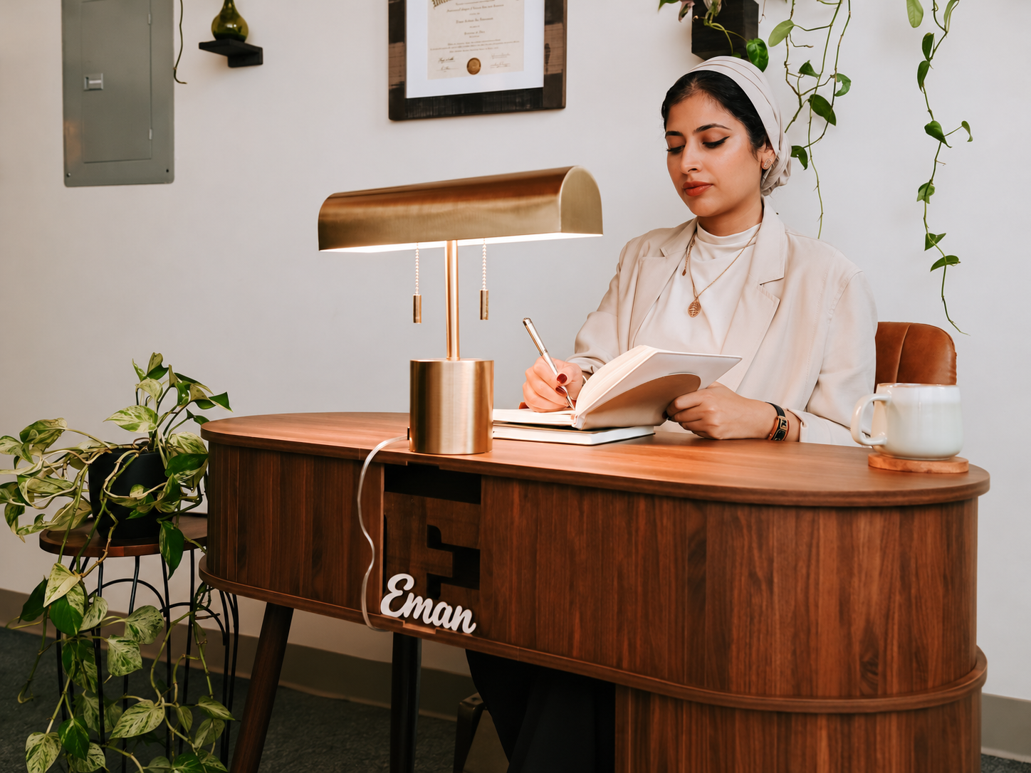 A woman with dark hair tied back, wearing a white blazer and headscarf, writing in a notebook at a wooden desk with a gold desk lamp, a white mug, and plants around her, with a diploma framed on the wall behind her.