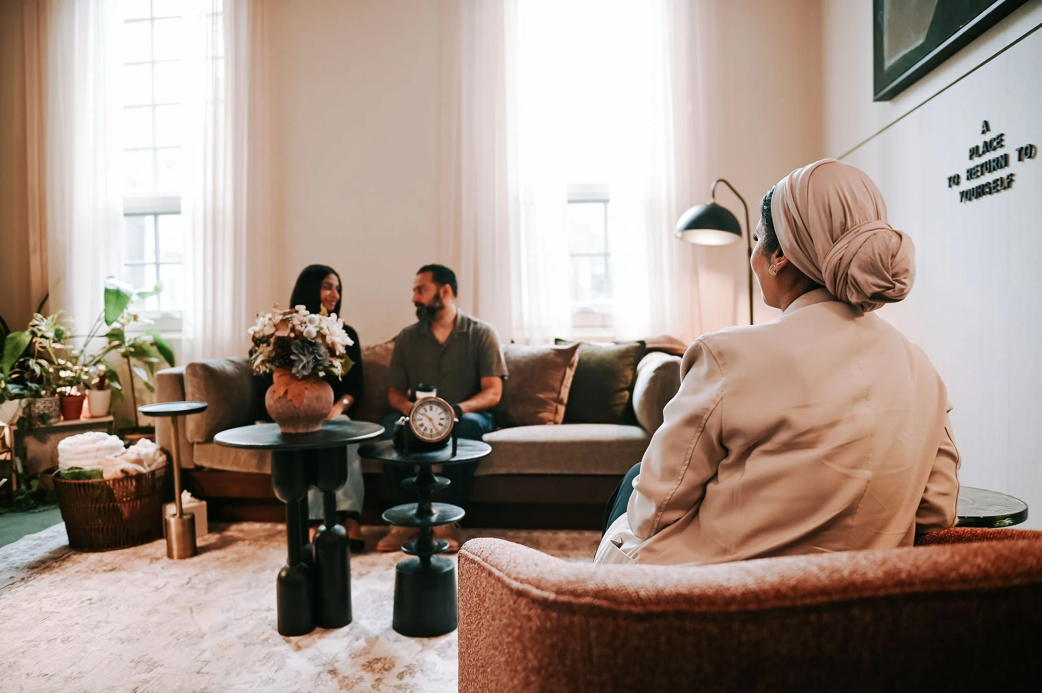 A woman wearing a peach-colored headscarf sitting in the foreground, facing away. In the background, a man and a woman are sitting on a couch, smiling and holding a bouquet of flowers in a cozy, well-lit living room.