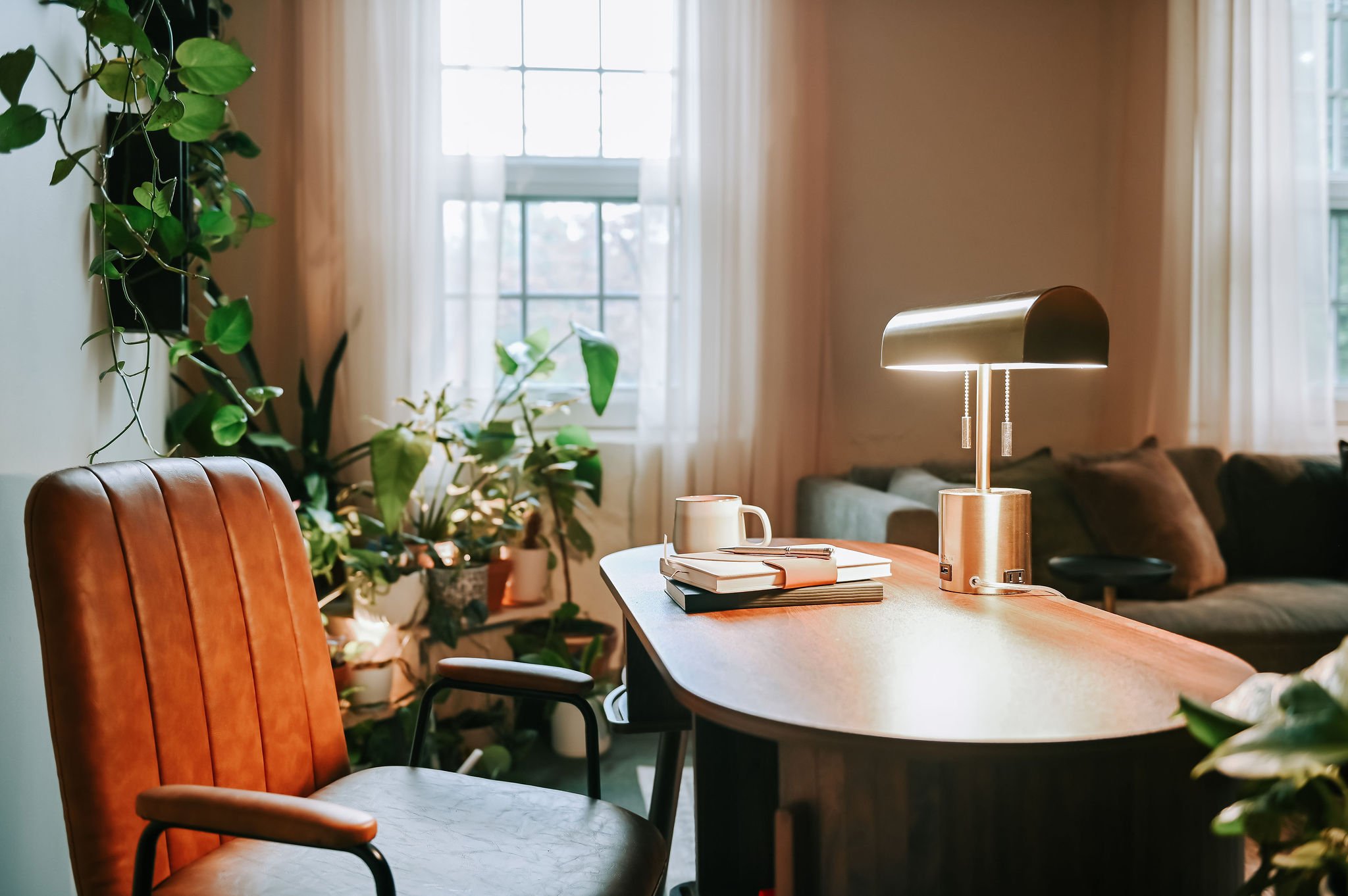 Interior of a cozy room with a wooden table, an orange leather chair, a black desk lamp, a white mug, a stack of notebooks, and several green houseplants near large windows with white curtains.