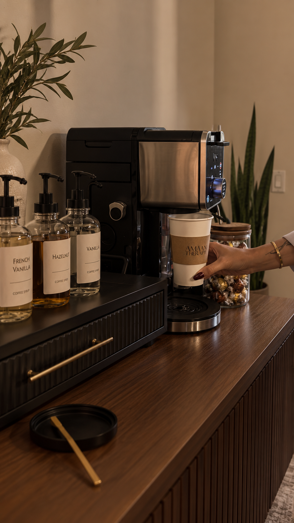 A person is holding a paper coffee cup under a coffee machine that is dispensing coffee. On the counter, there are bottles labeled with various flavored syrups, a large jar of wrapped candies, a black ashtray with a wooden stir stick, and a potted pl