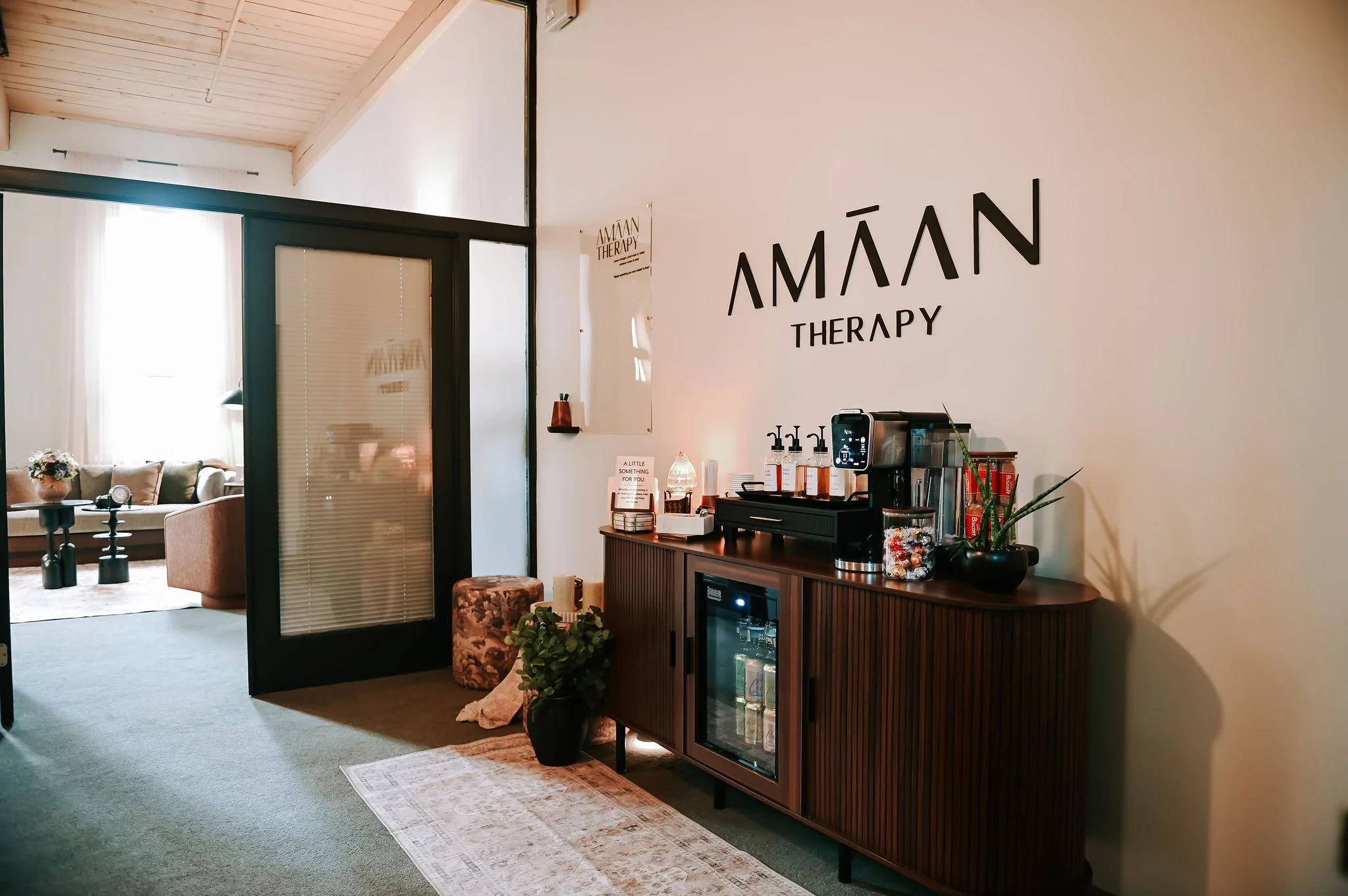 Interior of a wellness or therapy center with the words 'AMÁN THERAPY' on the wall, a counter with drinks and decorations, and a seating area visible through a glass door with sheer curtains.