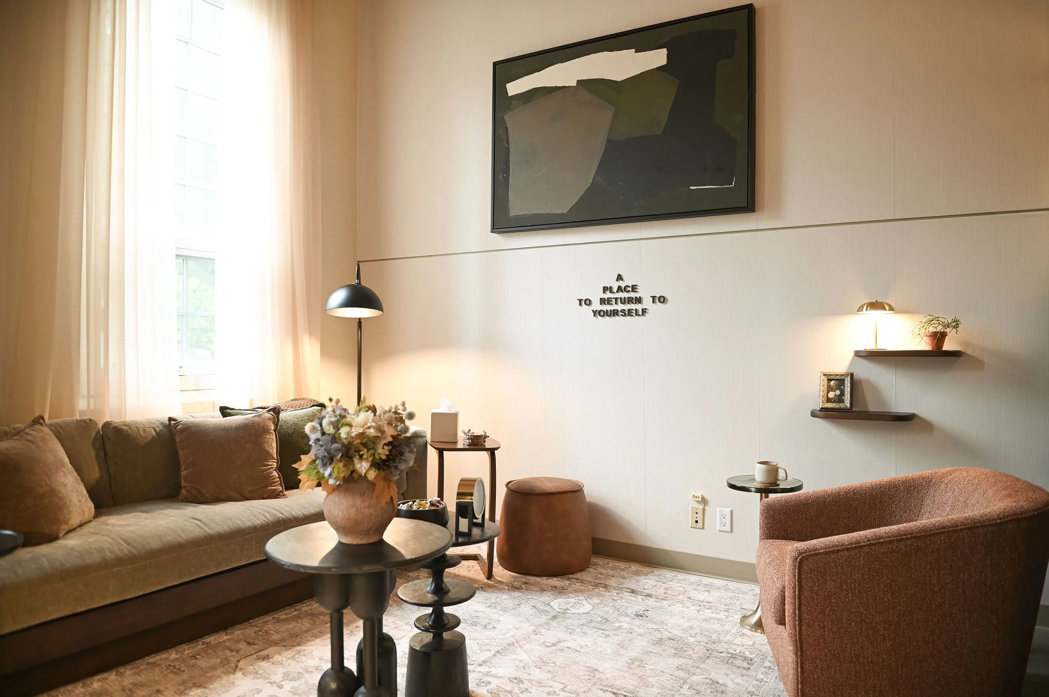A cozy living room with beige sofa, brown cushions, a black coffee table with a flower vase, a side table with a cup, wall art, lamps, and a small shelf.